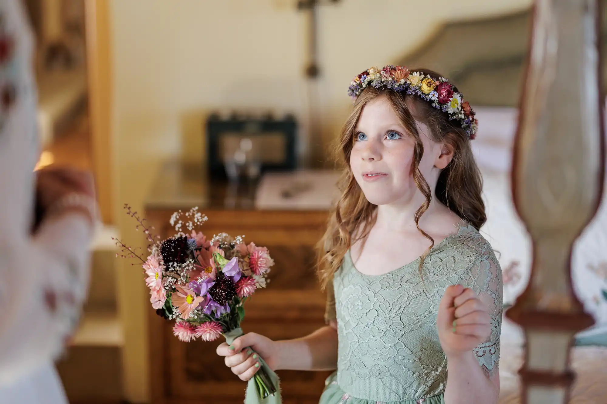 A young girl with wavy light brown hair wears a floral crown and a green lace dress. She smiles and holds a bouquet of colourful flowers, standing indoors near a wooden chest of drawers and a bed, in a softly lit, cosy room.