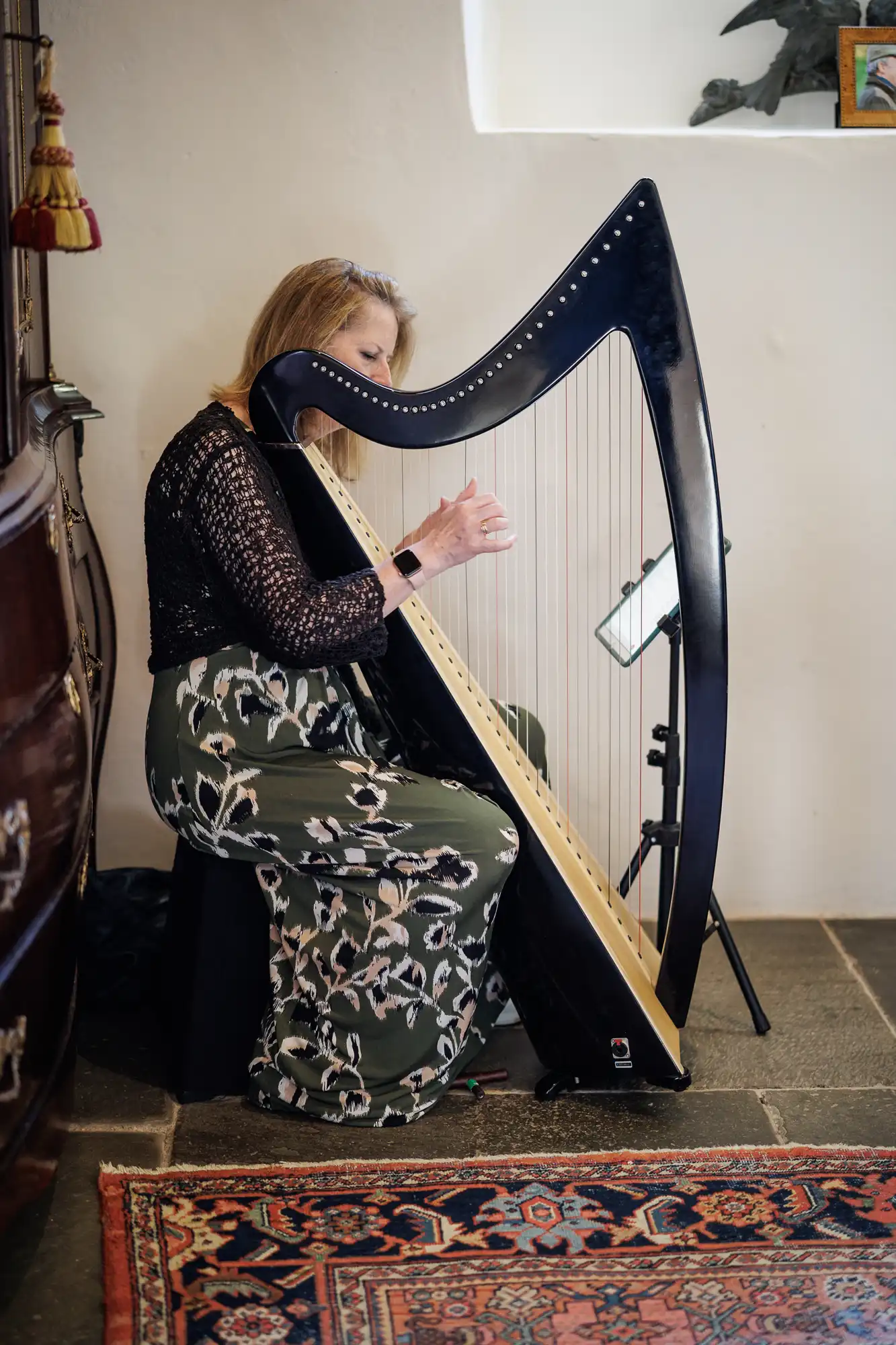 A woman with blonde hair, wearing a black lace top and green patterned skirt, sits on a stool playing a large black harp in a cosy room with white walls, stone floor, and a patterned rug. A music stand and lamp are nearby.