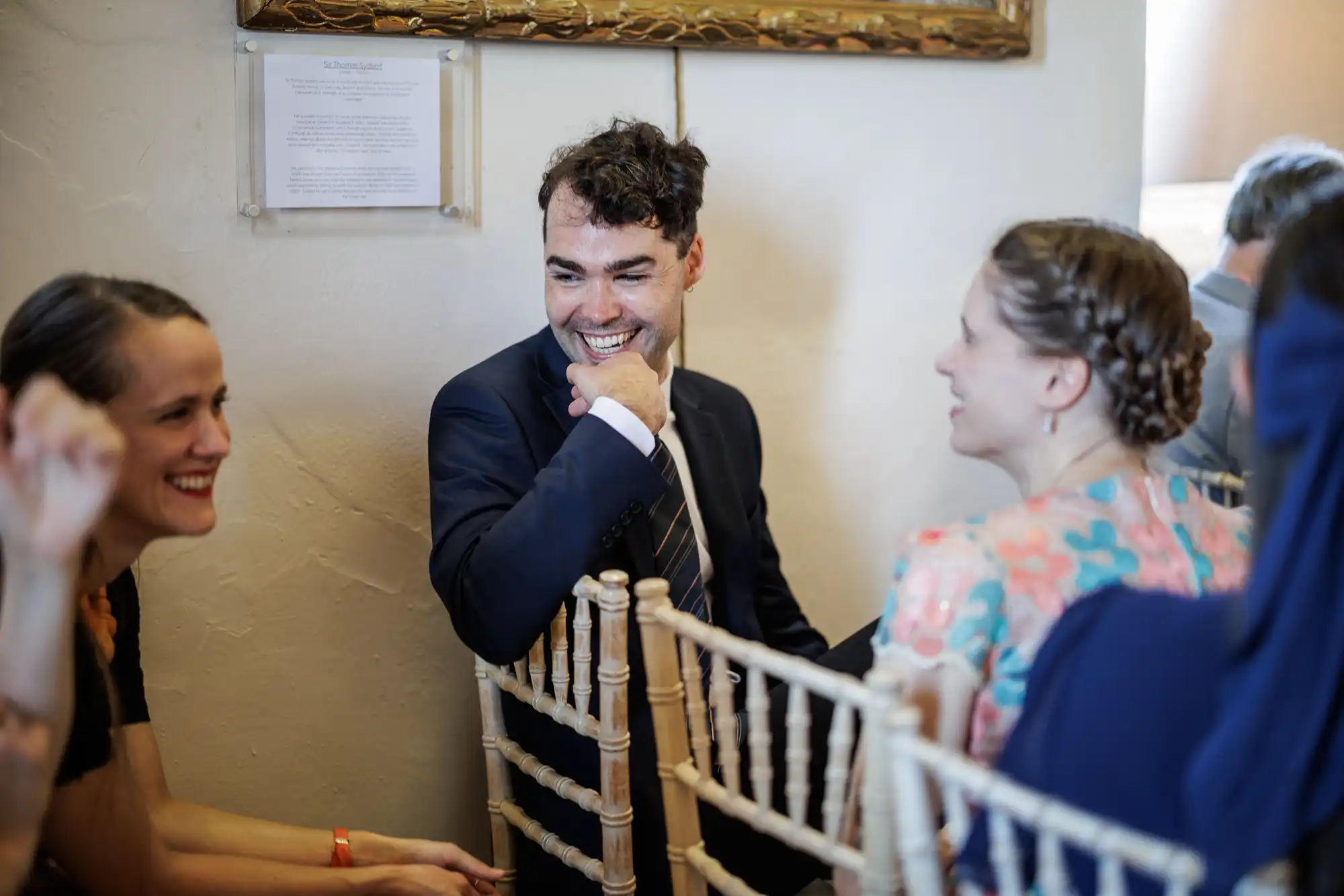 A man in a dark suit and striped tie sits among others on light wooden chairs, smiling and resting his chin on his hand. Two women near him, one in a floral dress, are also smiling, suggesting a joyful conversation at a formal event.