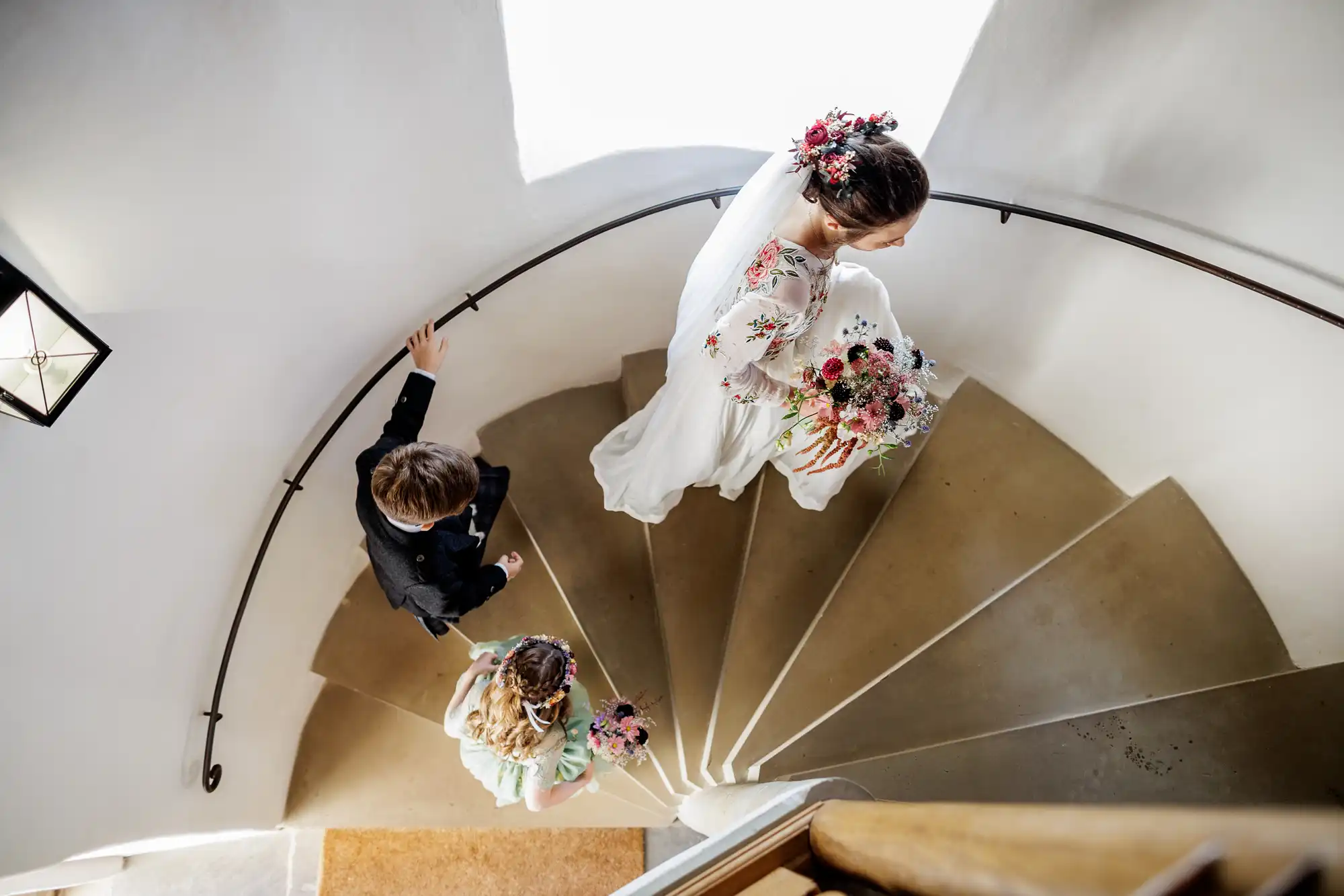 Overhead view of a bride in a white dress with floral hair accessories and bouquet, walking up a curved staircase, followed by a young girl holding flowers and a boy in formal attire, all bathed in soft natural light from a nearby window.