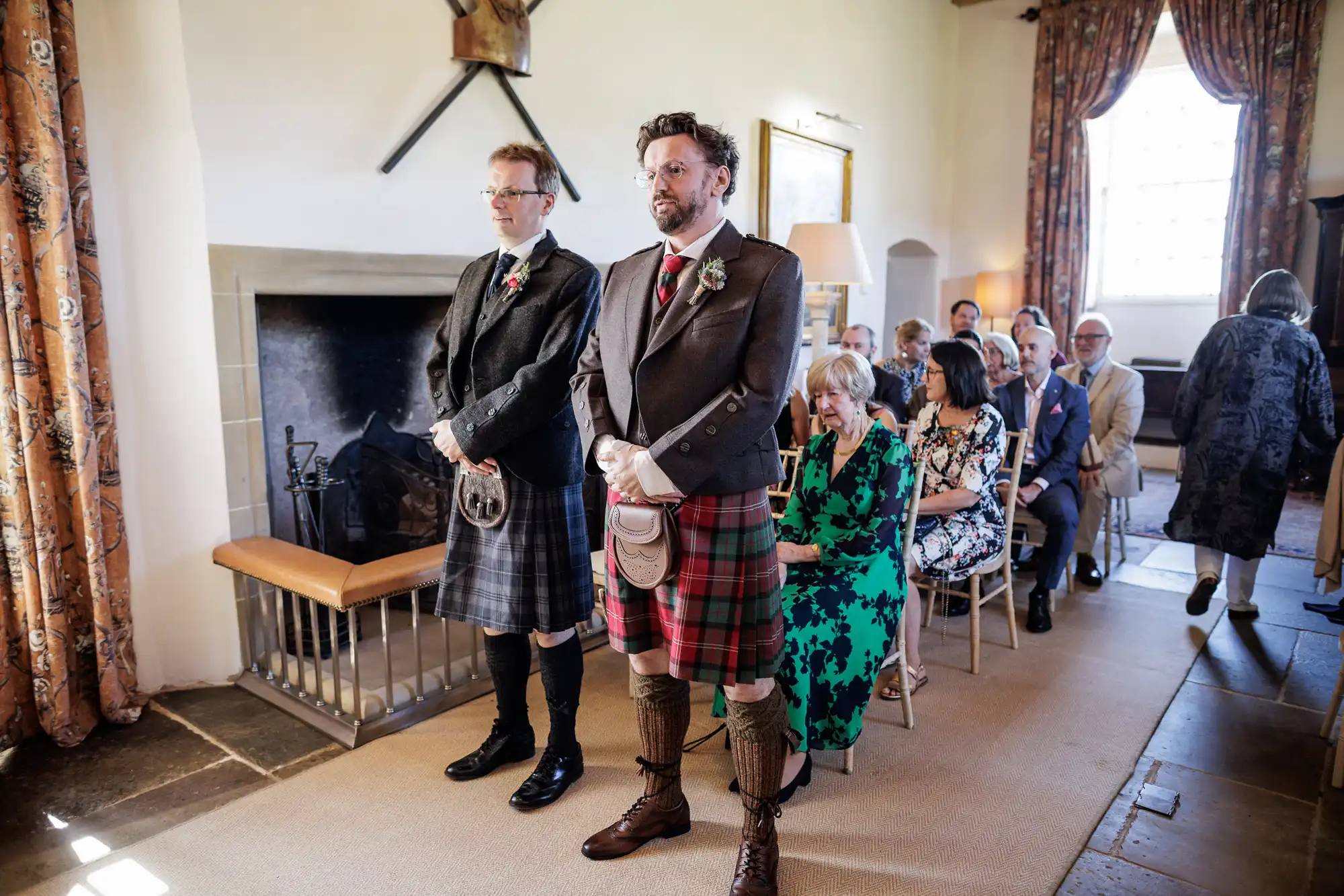 Two men in kilts stand at the front of a small, sunlit room with guests seated behind them. The men wear traditional Scottish attire; one in muted colours, the other in red tartan. The room has patterned curtains, a fireplace, and wooden details.