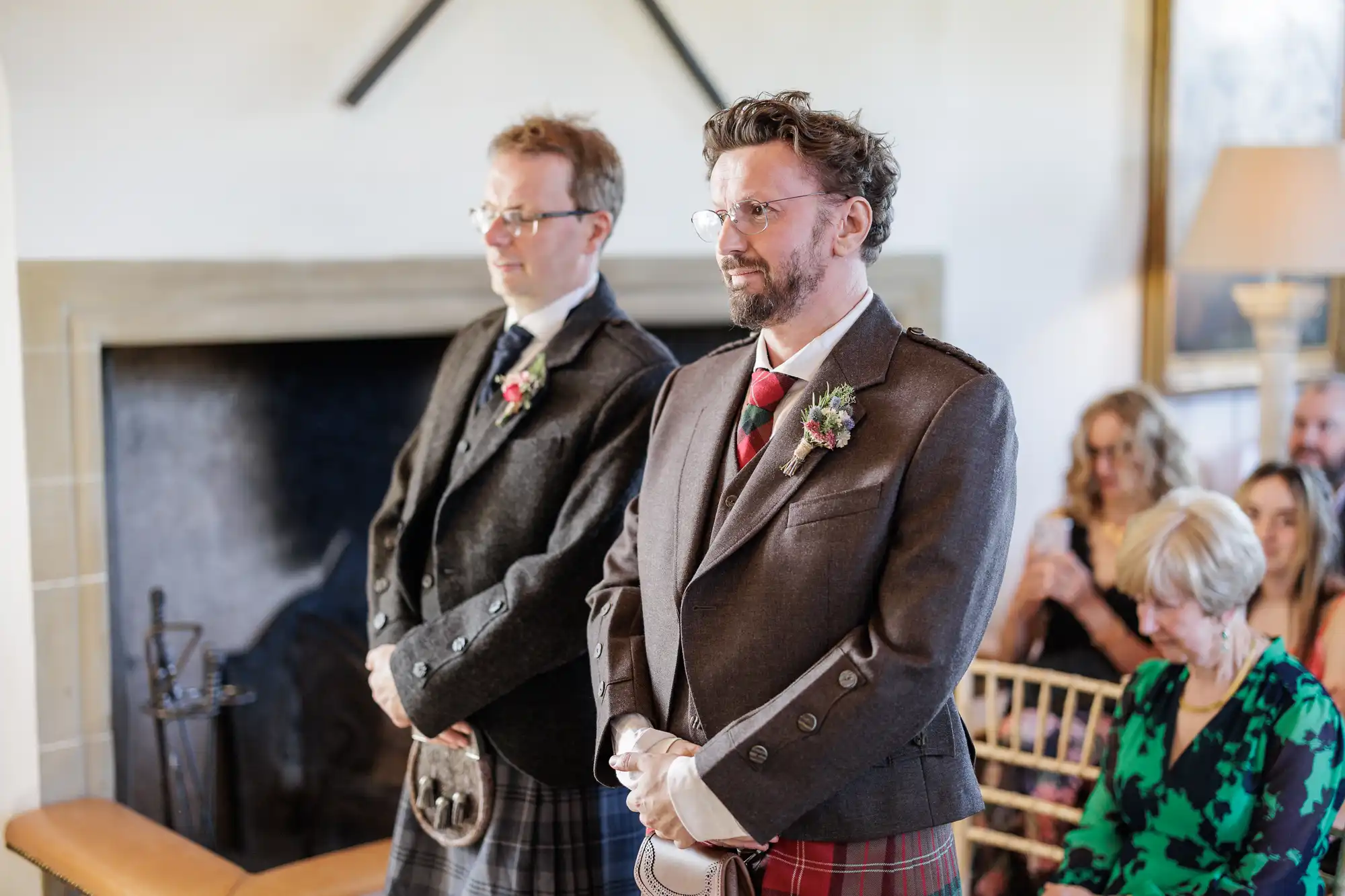 Two men in kilts and formal jackets stand side by side, smiling at the front of a room. Both wear buttonholes. Guests sit in rows behind them, some blurred, watching attentively. A fireplace and wall décor are visible in the background.