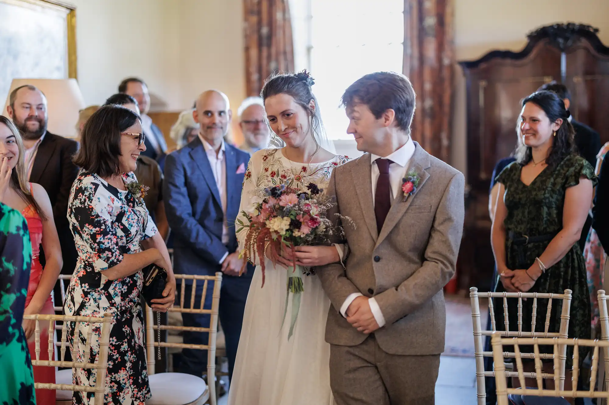 A bride in a white dress holding a bouquet stands beside a groom in a brown suit. They smile at each other, surrounded by seated and standing guests in formal attire inside a light-filled, elegant room.