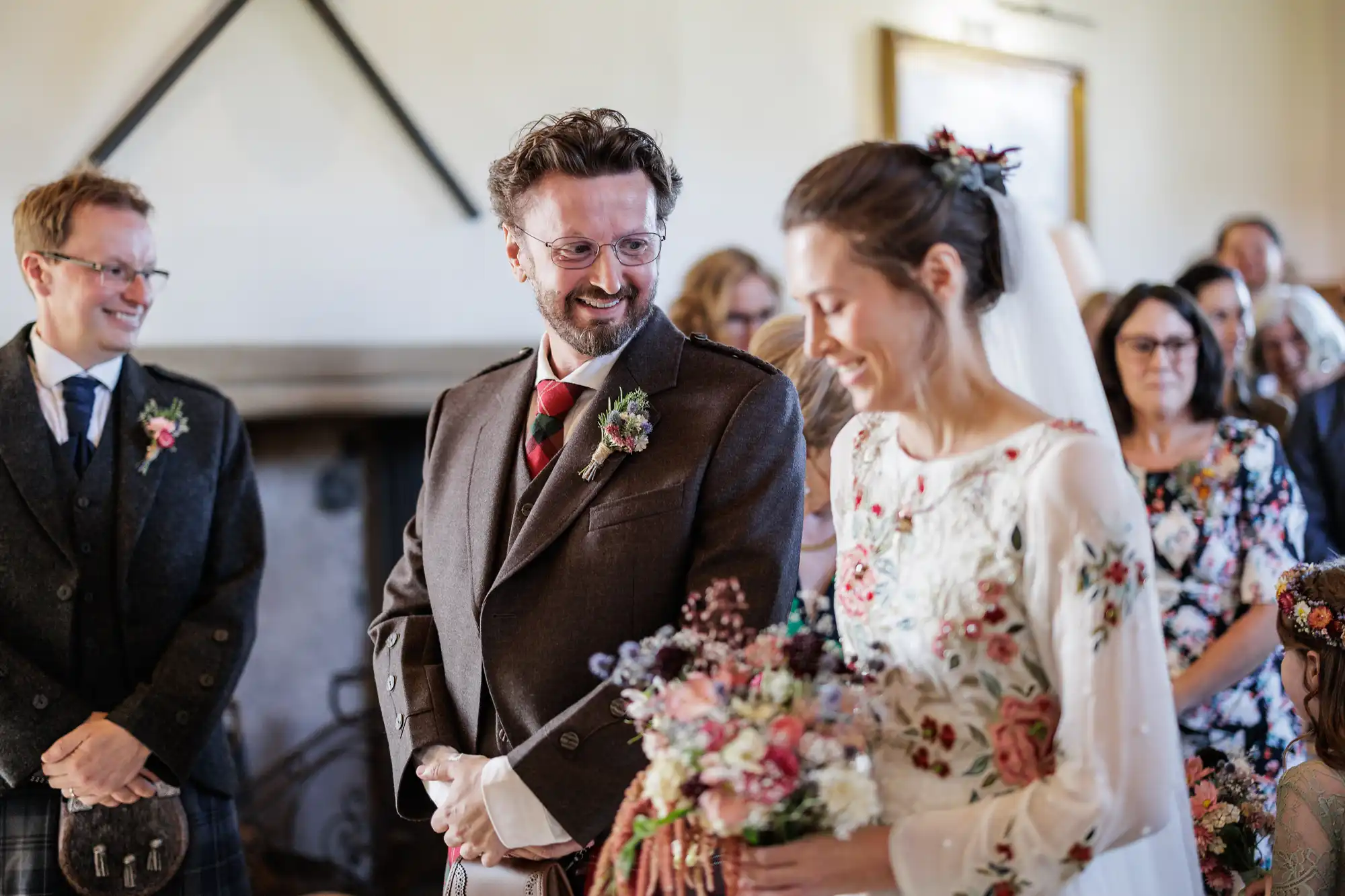 A groom in a brown suit smiles at a bride in a white floral dress holding a bouquet. Guests, some wearing glasses and floral attire, watch happily in the background. The setting is indoors with warm light and a visible fireplace.
