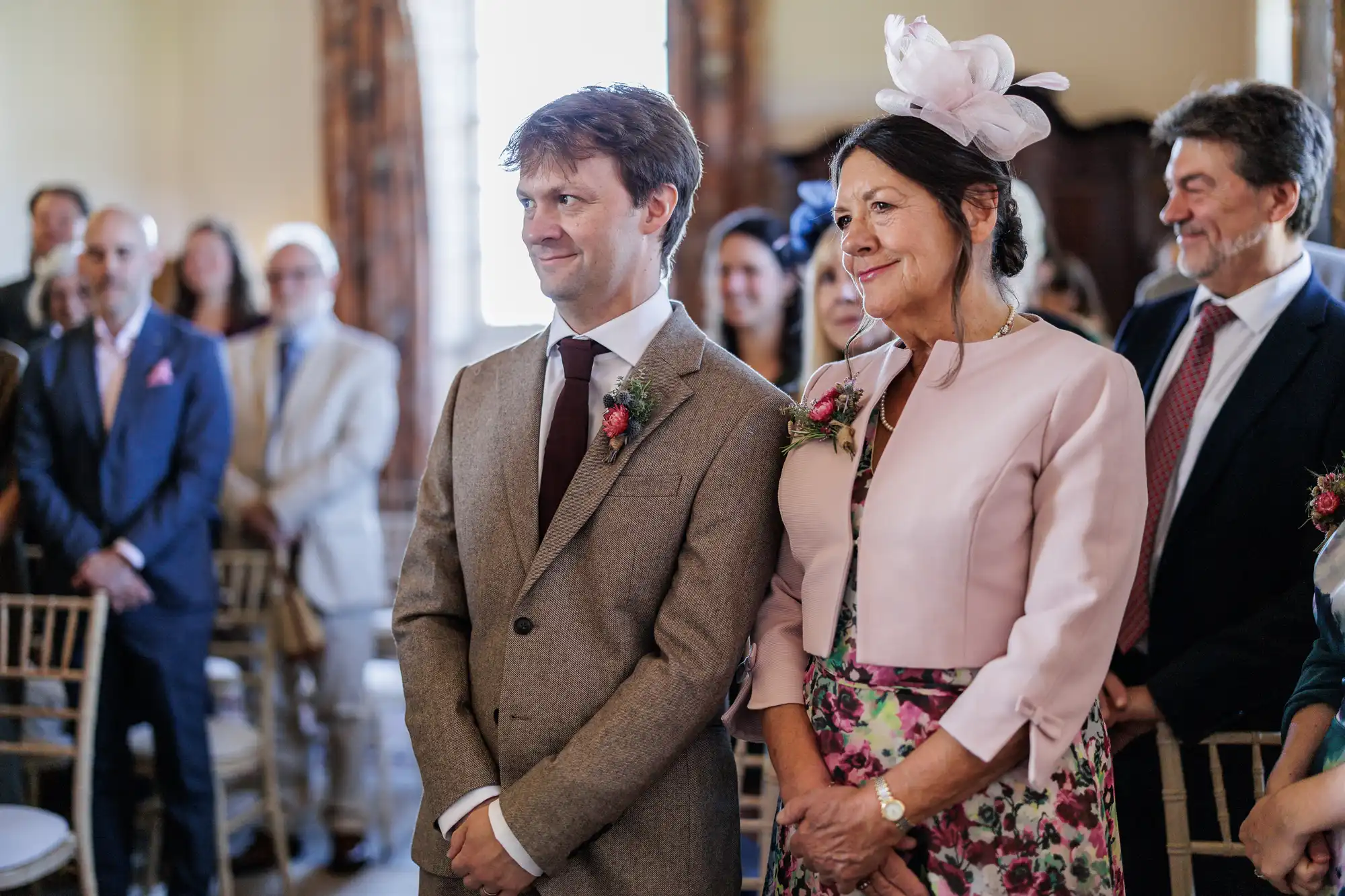 A smiling man in a brown suit and burgundy tie stands beside a woman in a light pink jacket, floral dress, and pink fascinator at a formal indoor gathering, likely a wedding. Several people in formal clothing are seated and standing behind them.