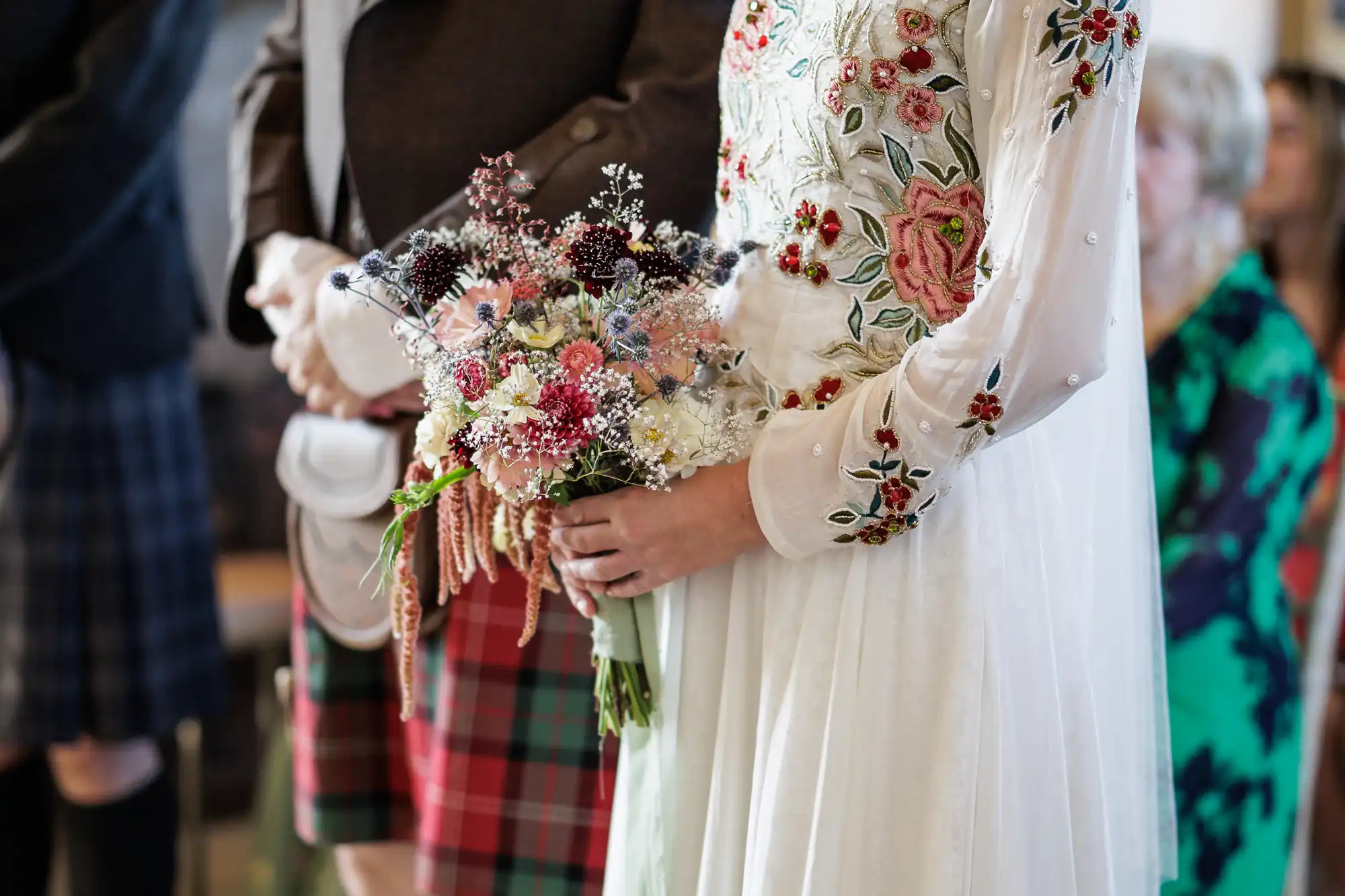 A bride in a white dress with colourful floral embroidery holds a bouquet of mixed flowers beside a groom in Scottish attire, including a brown jacket and red tartan kilt. Guests in colourful outfits are blurred in the background.