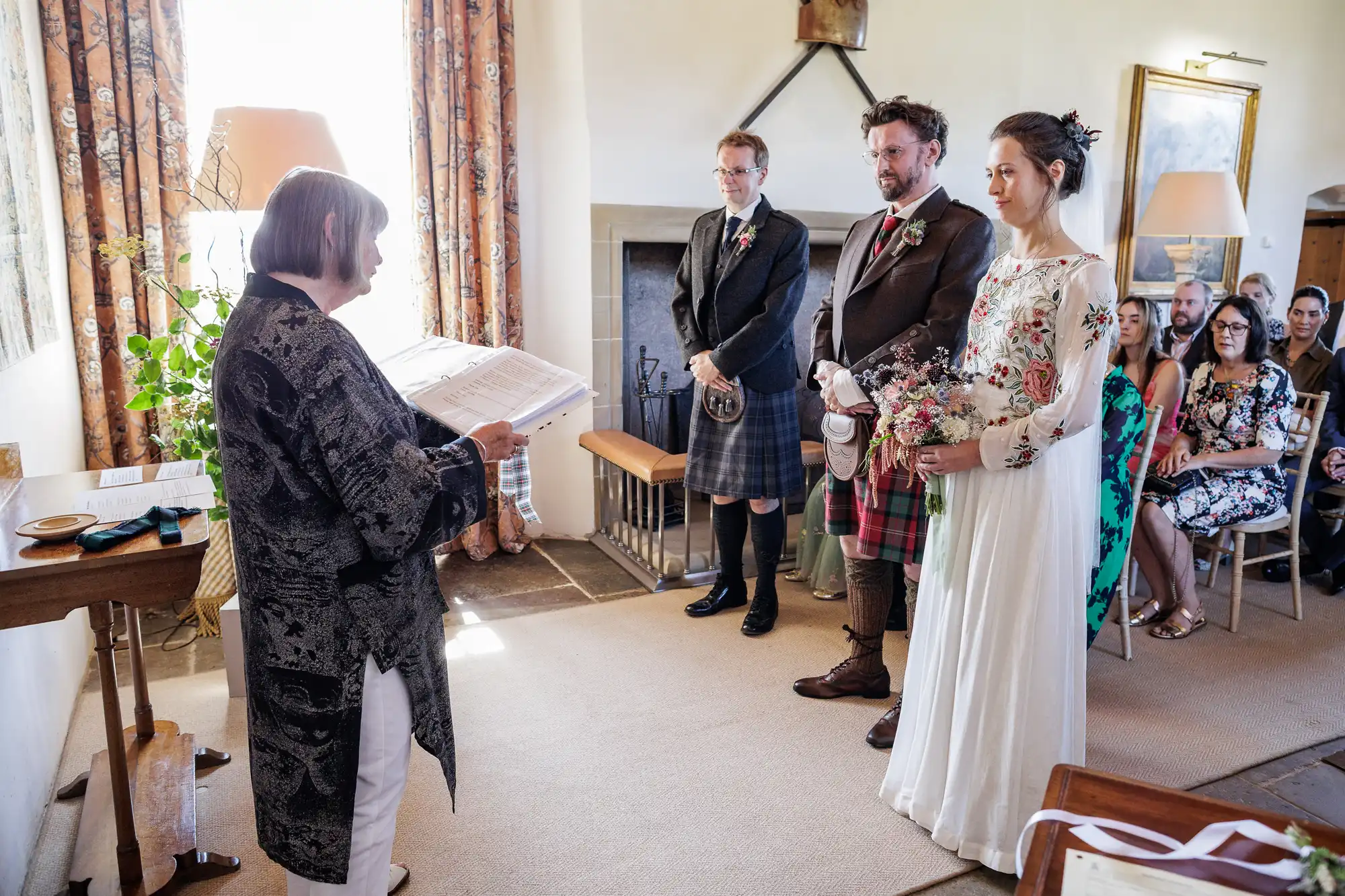A wedding ceremony in a cosy room: an officiant reads from papers to a bride in a white dress with floral embroidery and a groom in a checked kilt. A man in a dark kilt stands beside them. Seated guests watch, sunlight streaming through patterned curtains.