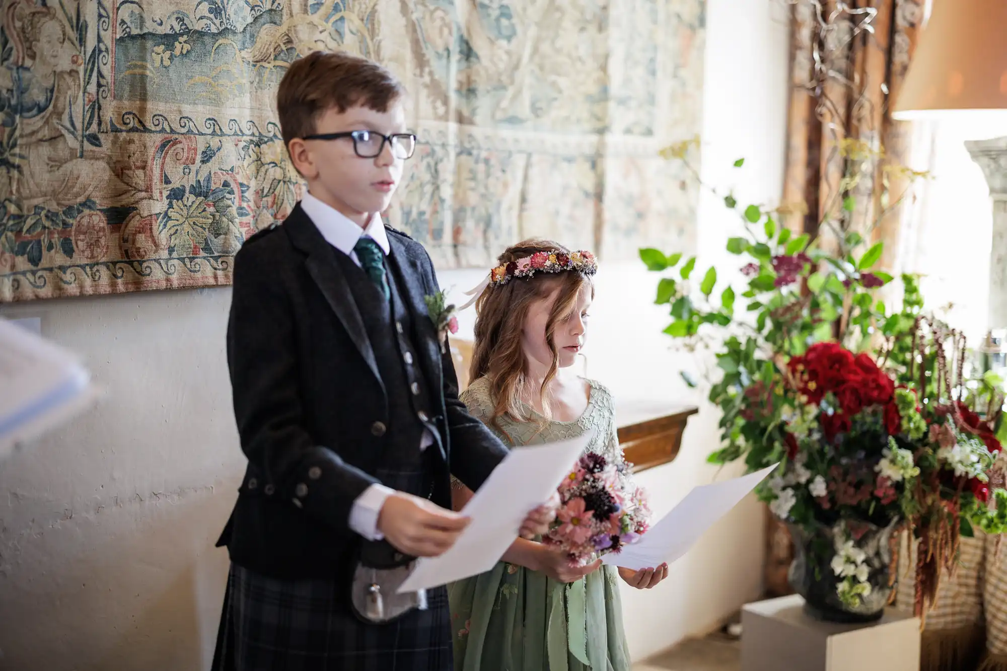 A young boy in glasses and a suit holds papers while standing beside a girl in a green dress and flower crown holding a bouquet. They appear to be reading or presenting at a formal indoor event, with floral arrangements and a tapestry in the background.