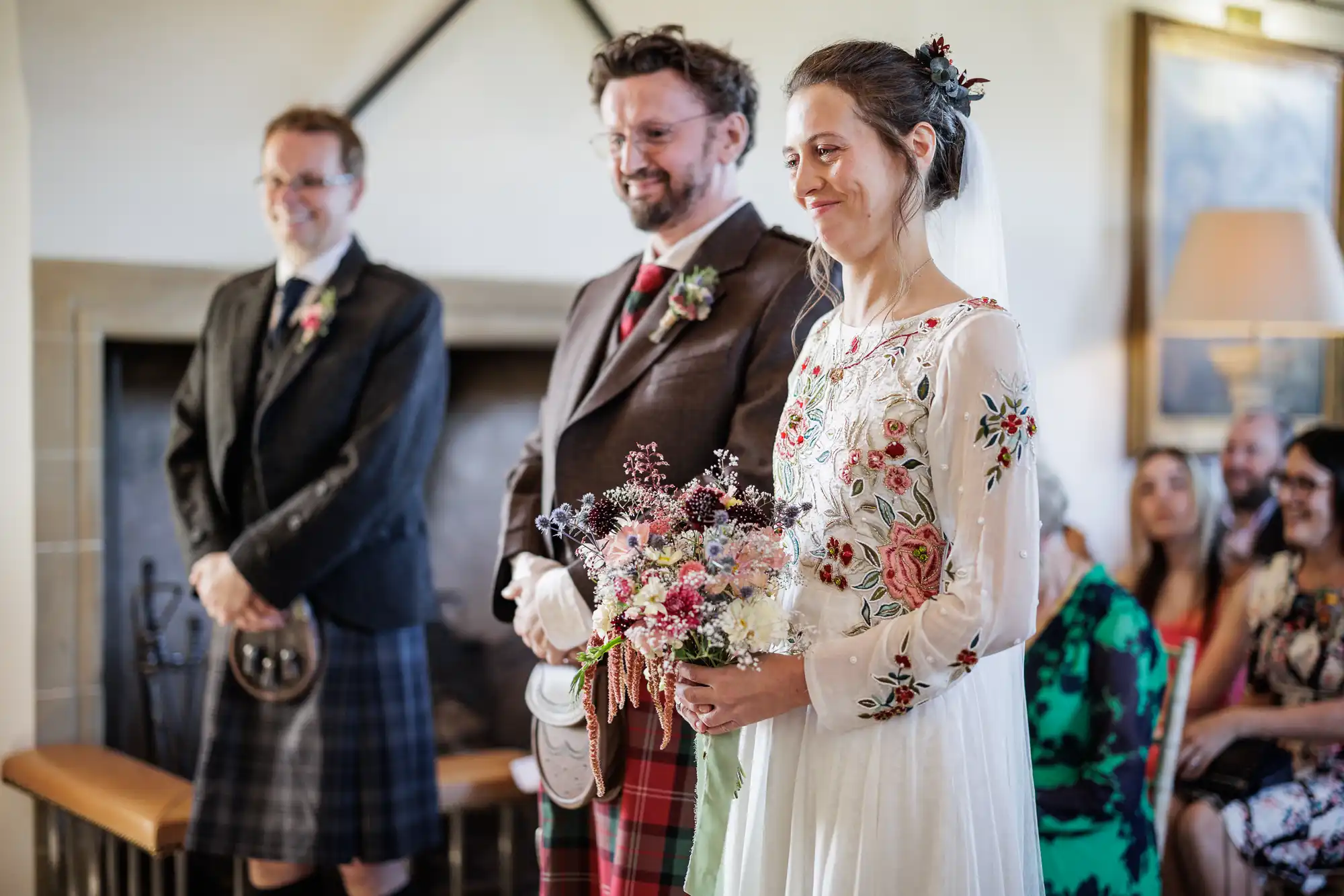 A bride in a white, floral-embroidered dress holds a colourful bouquet and smiles beside a groom in a tartan kilt and jacket. Another man in a dark jacket and kilt stands nearby. Guests are seated in the background, and the setting is warm and softly lit.