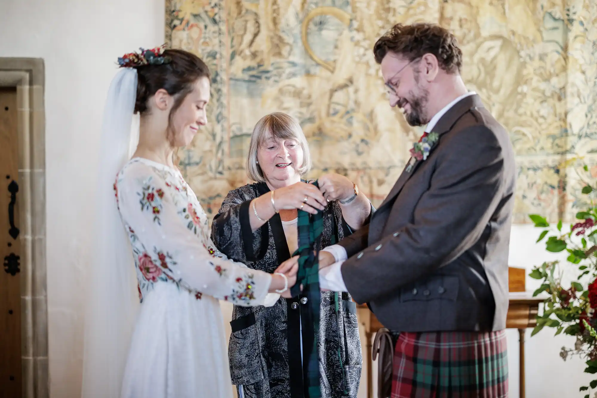 A smiling bride and groom stand facing each other, holding hands as an officiant ties a green and blue tartan ribbon around their joined hands during a wedding ceremony in a decorated indoor setting.