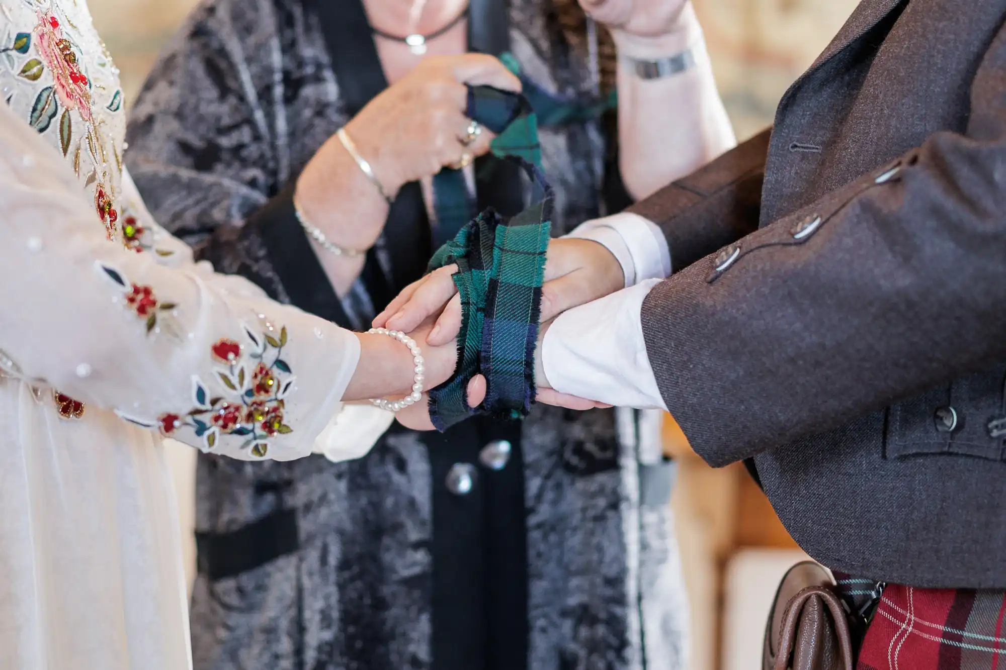Close-up of three people’s hands during a handfasting ceremony. Two people hold hands while a third wraps a green and blue tartan cloth around them. One participant wears embroidered white, another wears a dark jacket and kilt; the third wears a patterned jacket.
