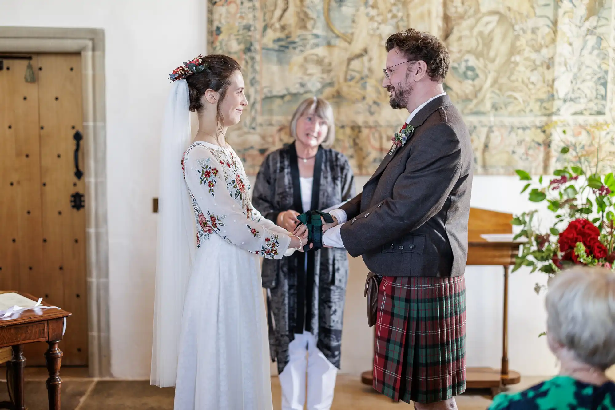 A bride in a white, long-sleeved floral dress and veil holds hands with a groom in a brown jacket and red tartan kilt. They smile at each other during a ceremony, while an officiant stands behind them, reading, with flowers and tapestry in the background.