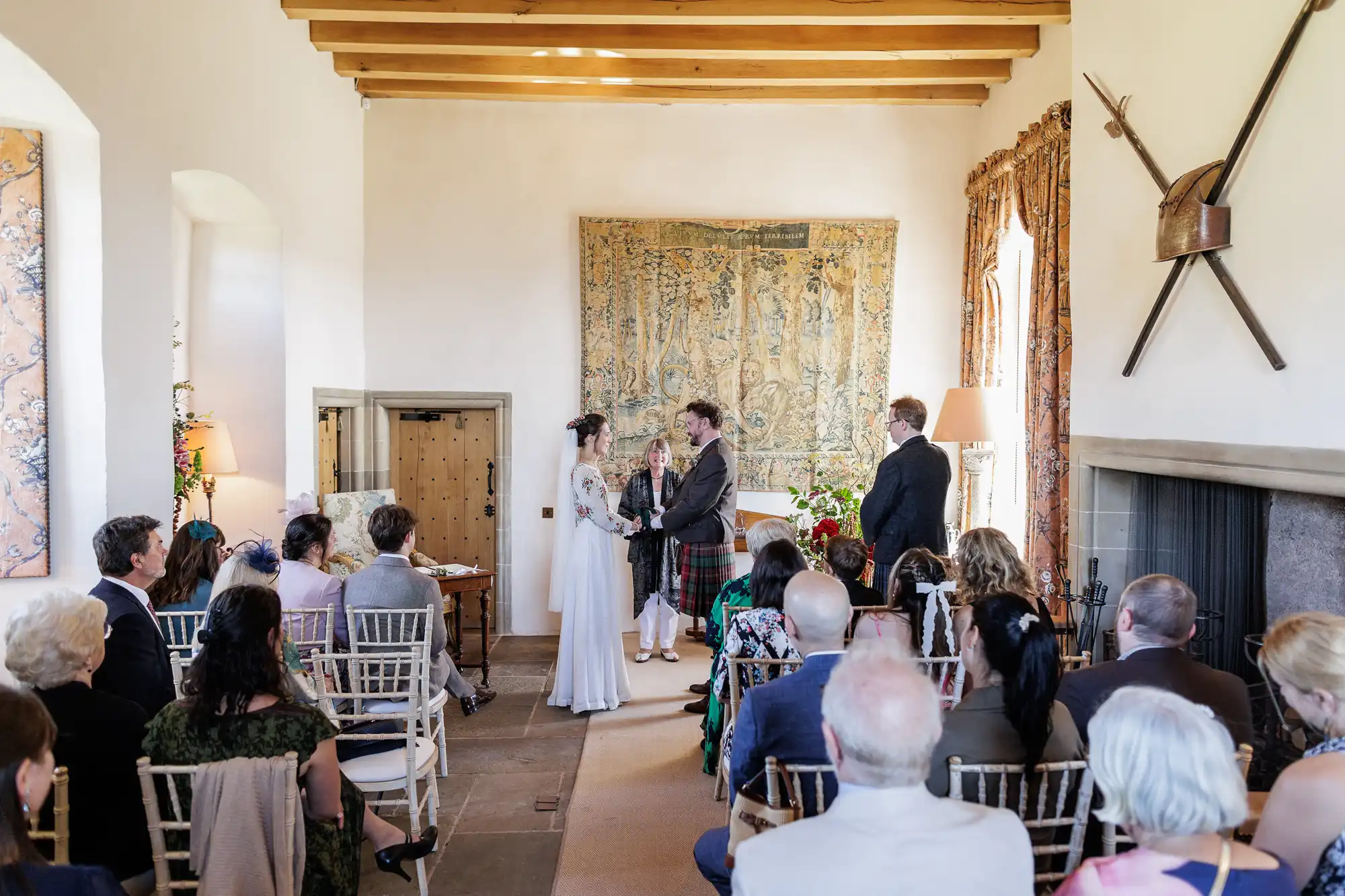 A couple stands facing each other holding hands, exchanging vows in a bright room with wooden beams and medieval decor. Guests are seated on white chairs, attentively watching. A tapestry and flowers decorate the space; sunlight streams through tall windows.