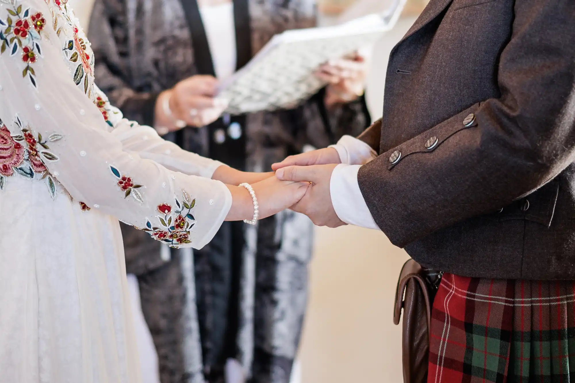 A close-up of two people holding hands at a wedding ceremony. One wears a white embroidered dress, the other a dark jacket and tartan kilt. An officiant in a patterned robe holds a book in the background. The scene conveys intimacy and commitment.