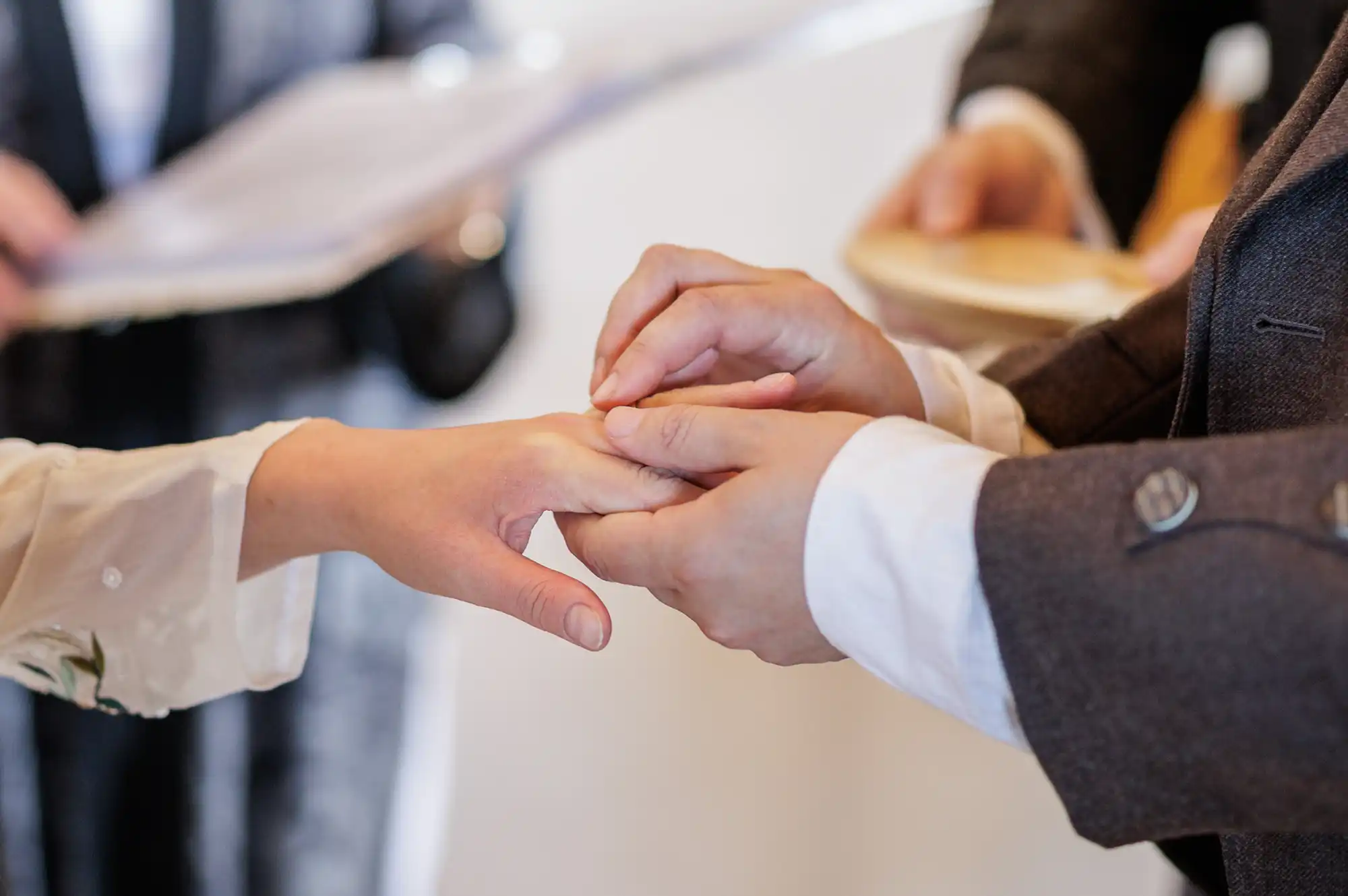 Close-up of one person placing a ring on another’s finger during a wedding ceremony. Both wear formal attire, with one in a dark suit and the other in light-coloured sleeves. A blurred figure in the background holds documents.