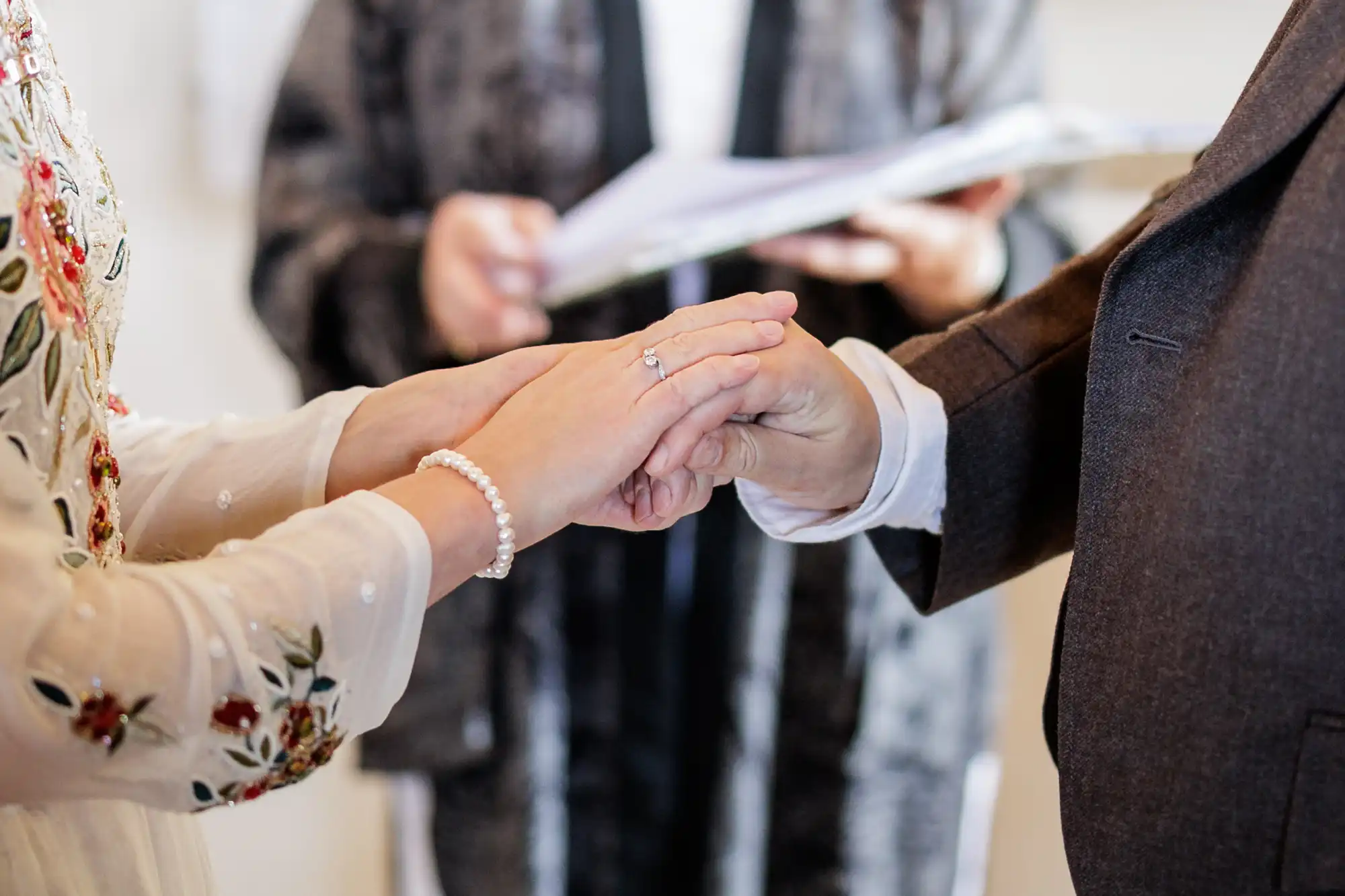 Close-up of two people holding hands during a wedding ceremony; one is wearing a white, floral-embroidered dress and a pearl bracelet, the other a dark suit. A third person in the background holds documents, slightly out of focus.