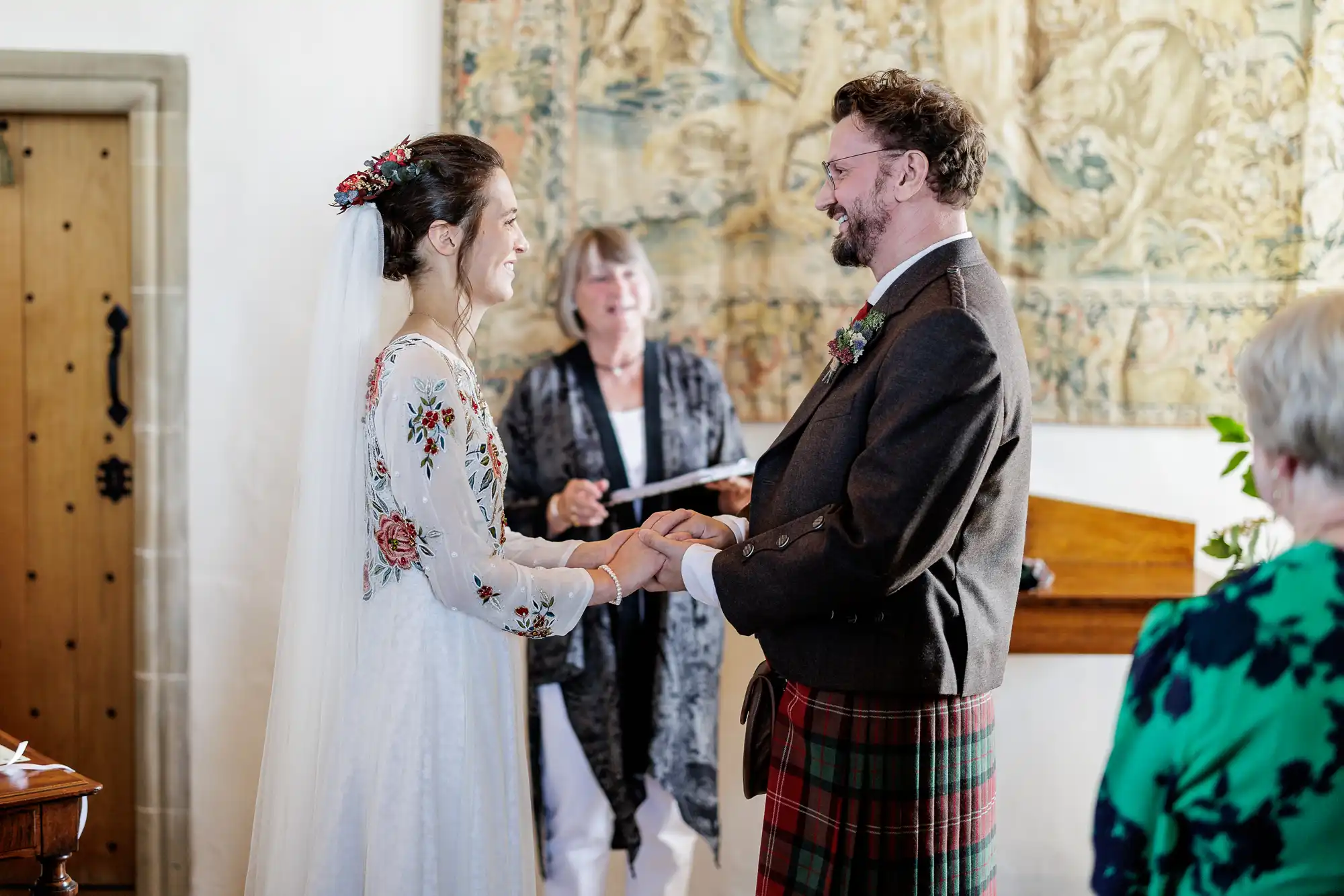 A bride in a white dress with floral embroidery and a groom in a dark jacket and tartan kilt exchange rings during a wedding ceremony. An officiant stands behind them, and a guest in green sits to the right. A tapestry hangs on the wall in the background.