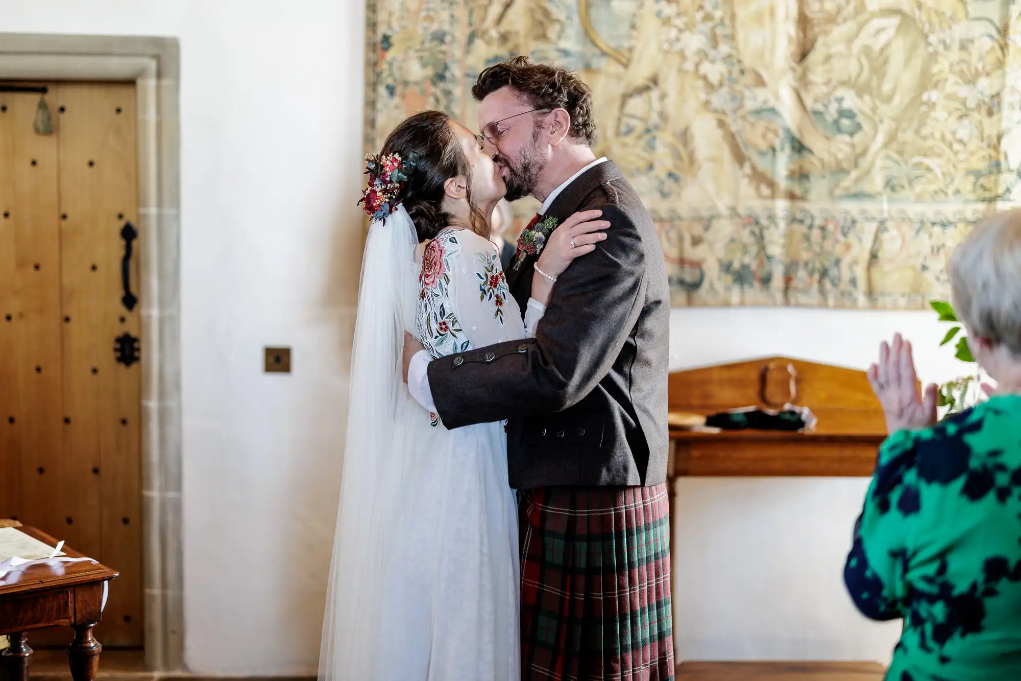 A bride and groom kiss indoors after their wedding ceremony. The bride wears a white dress with floral embroidery and a veil with flowers in her hair. The groom wears a brown jacket and tartan kilt. An older guest in green applauds in the foreground.