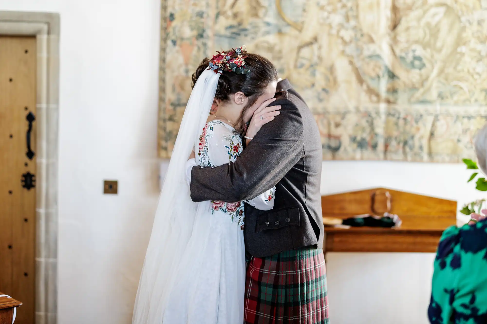 A bride in a white dress with floral embroidery and a groom in a brown jacket and checked kilt embrace closely, faces touching and partly hidden. The bride wears a veil and floral headpiece. Behind them is a tapestry and wooden bench in a softly lit room.