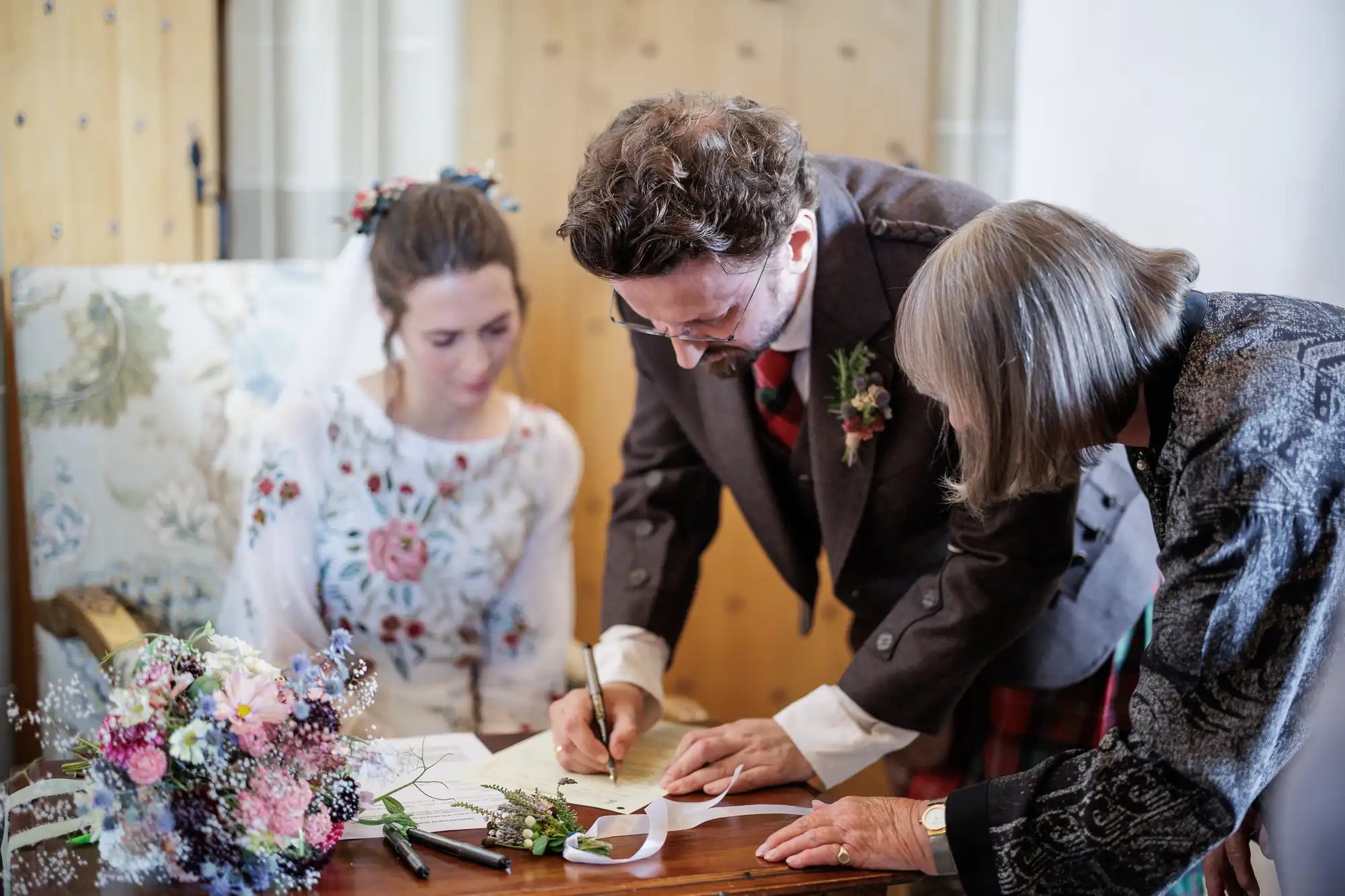 A bride in a floral dress and a groom in a dark suit sign wedding documents at a wooden table, assisted by an older woman. A bouquet of pastel flowers, pens, and greenery rest on the table. The scene is indoors, with soft, natural lighting.