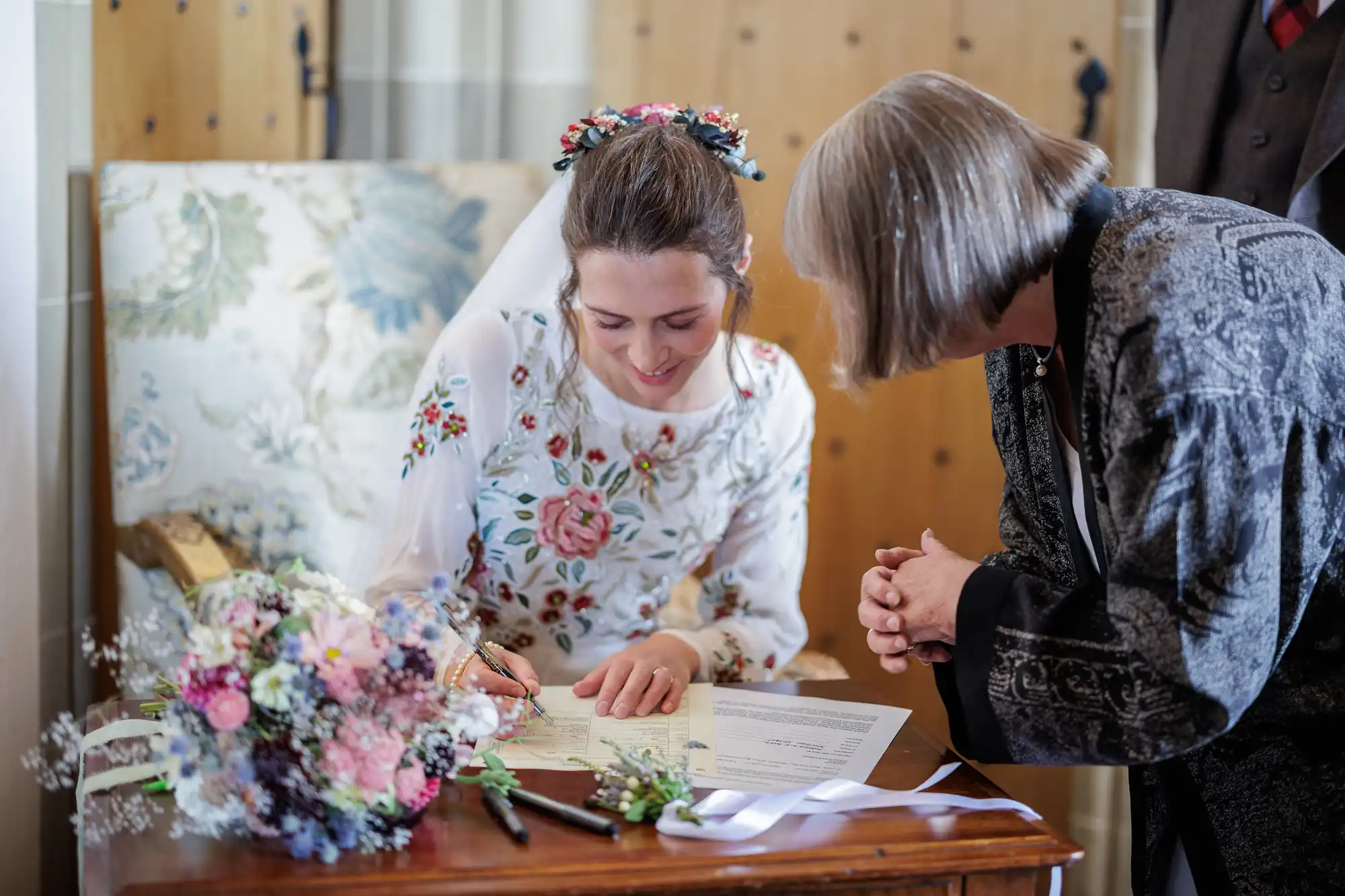 A smiling bride in a white, floral-embroidered dress signs a document at a wooden table, while a woman with short grey hair watches. A colourful bouquet, pen, and papers are on the table. The setting looks formal, possibly a wedding ceremony.