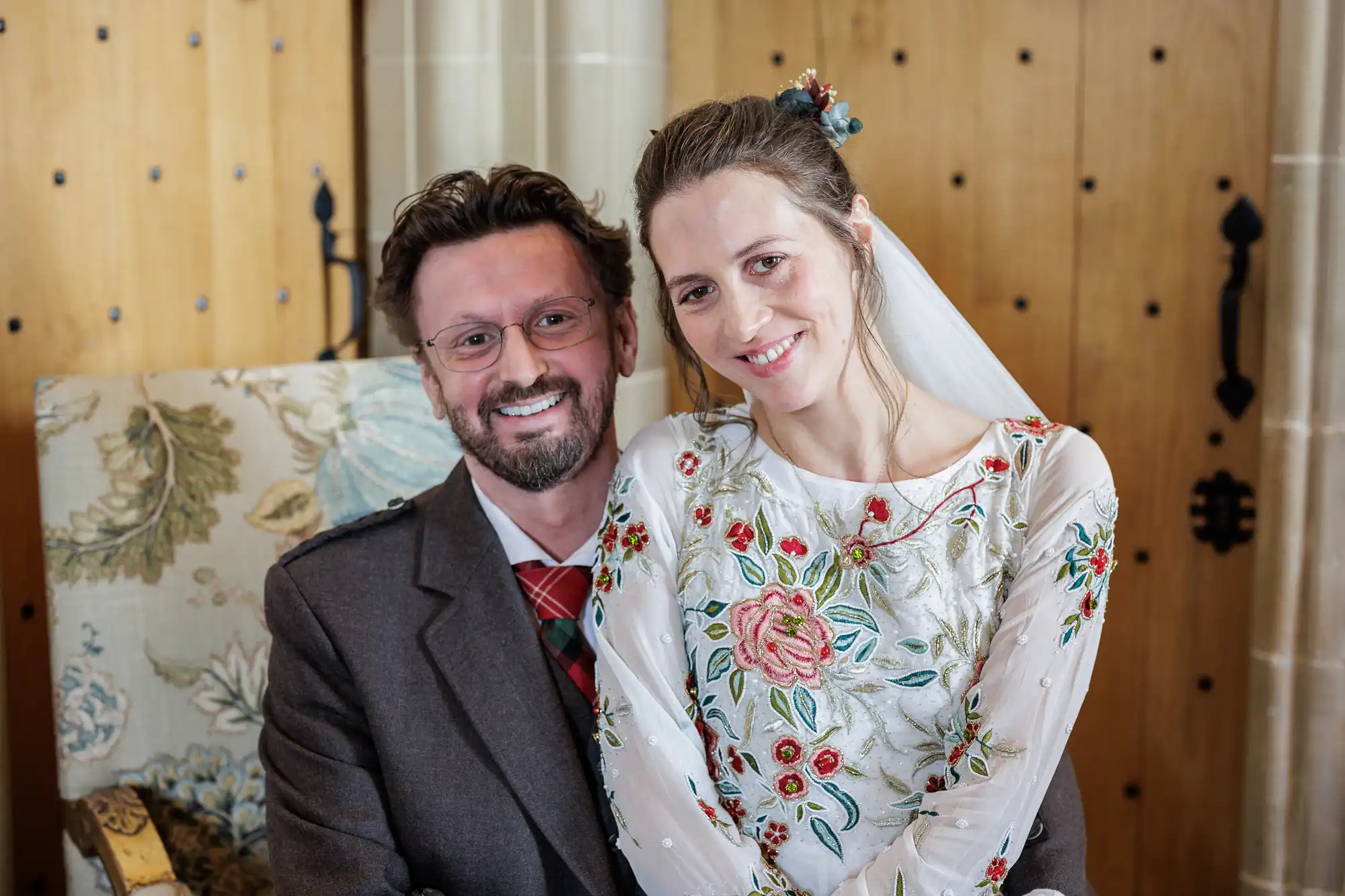 A smiling couple pose indoors. The man has glasses, short brown hair, and a beard, wearing a brown suit with a red tartan tie. The woman has light brown hair pulled back, wearing a white dress with colourful floral embroidery and a white veil.