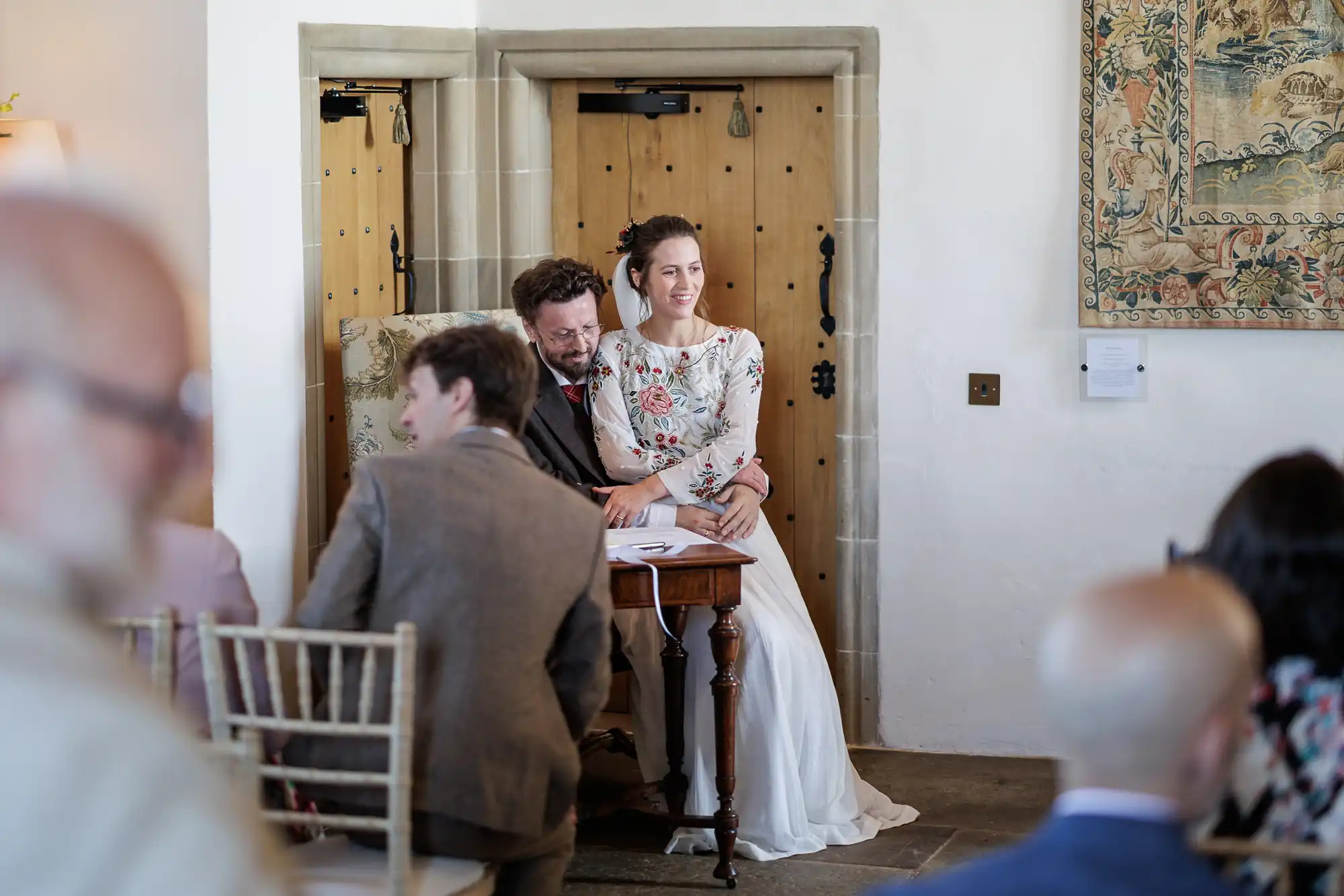 A bride in a white floral dress sits on a groom’s lap at a small table, both smiling. They are indoors by wooden doors. Guests in formal attire sit and stand in the foreground, some blurred, creating a candid, joyful atmosphere.