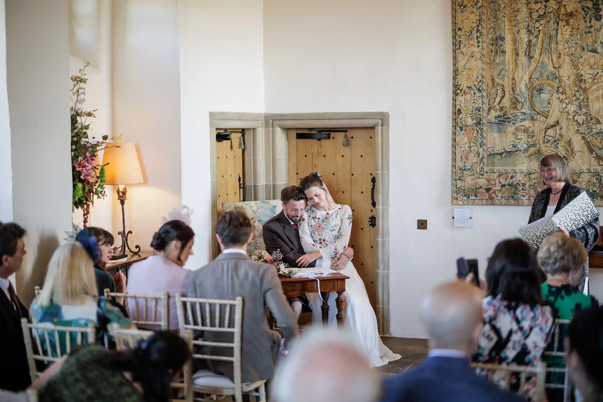 A bride in a white dress leans affectionately on a groom in a suit as they sit at a wooden table during a wedding ceremony. Guests are seated and watching, whilst a woman stands nearby holding a large present. The room is decorated with flowers and a large tapestry.