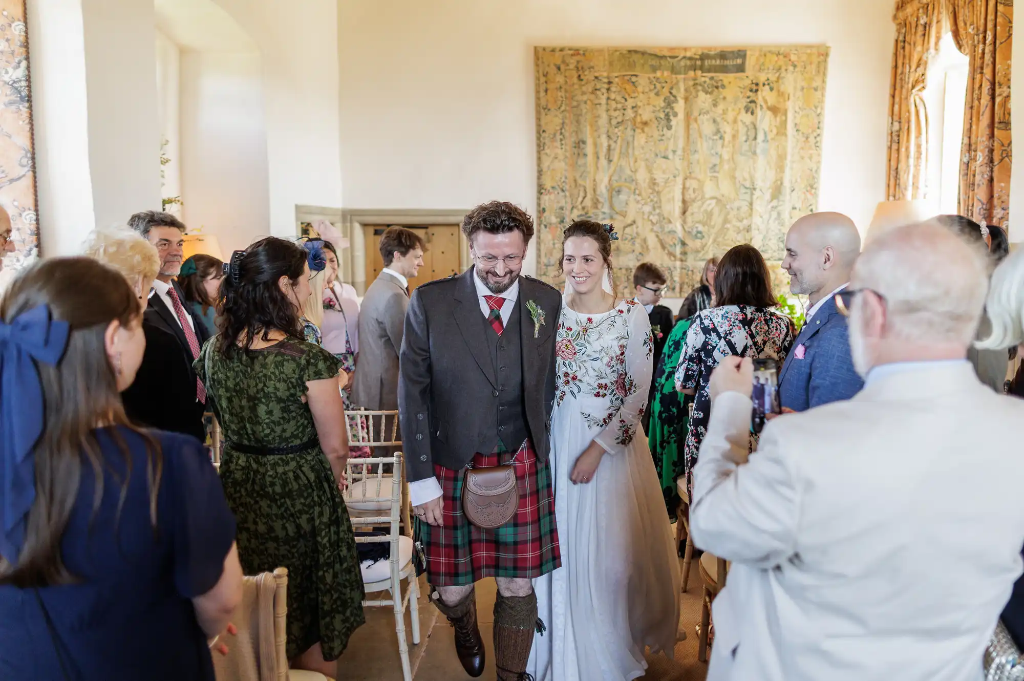 A smiling couple, the groom in a tartan kilt and jacket, and the bride in a long-sleeved white gown with floral embroidery, walks down an aisle in a bright room with guests standing and watching, some taking photos.