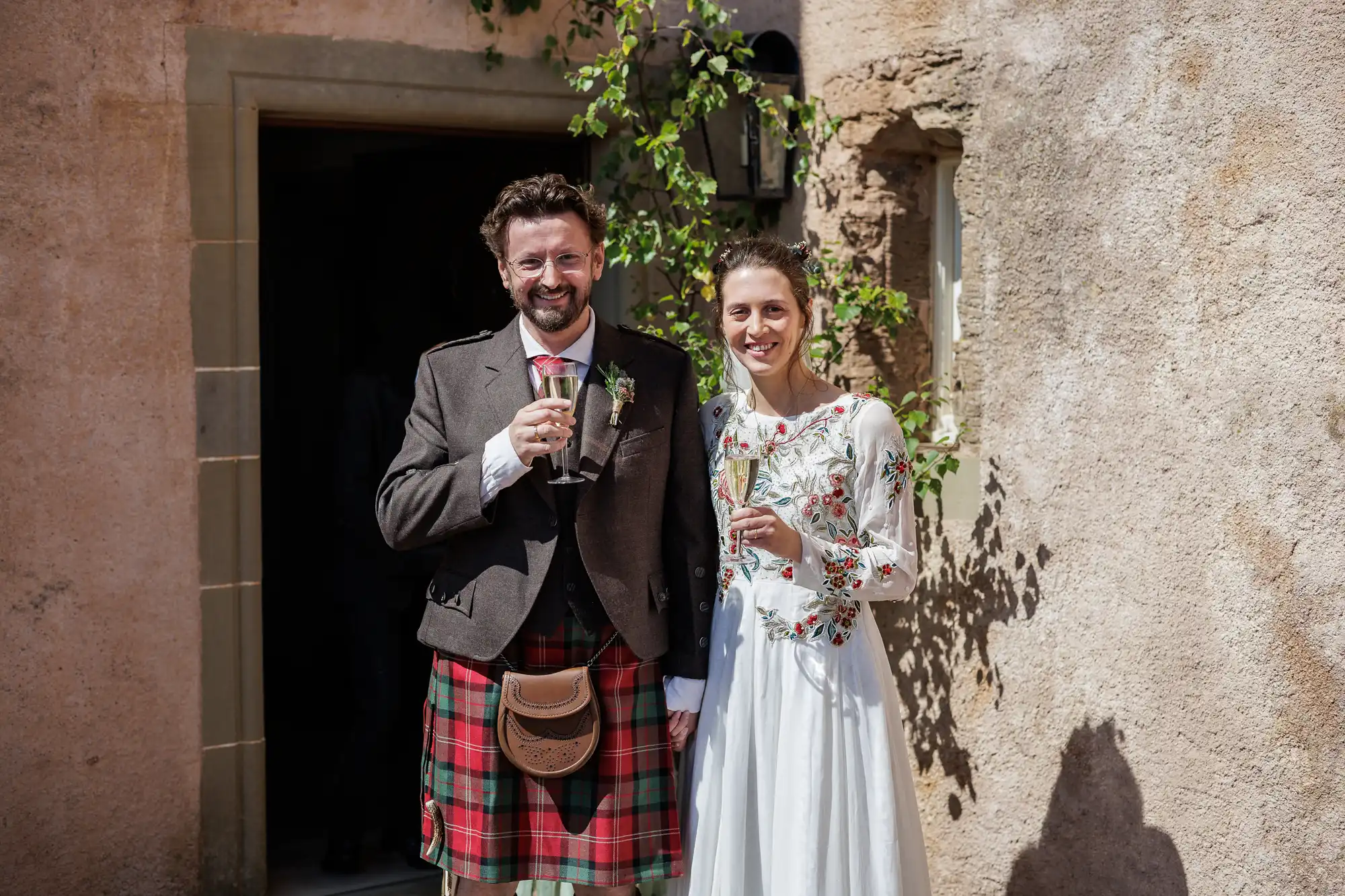 A smiling couple stands outside against a stone wall. The man wears a brown jacket, red tartan kilt, and sporran, holding a drink. The woman wears a white long-sleeve dress with floral embroidery, also holding a drink. Green vines grow nearby.