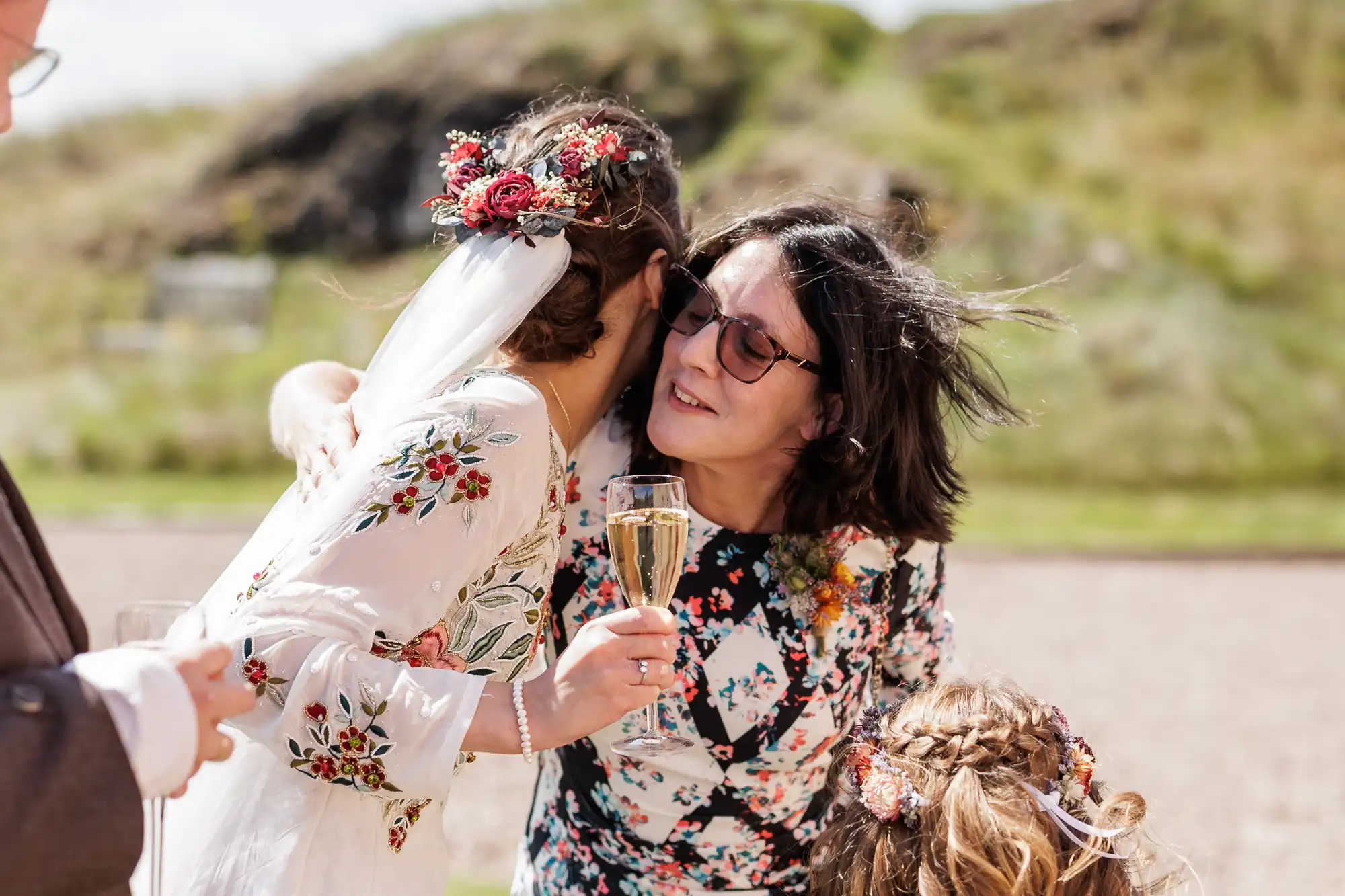 A bride in a floral dress and veil with red flowers hugs a woman in glasses holding a champagne flute. Both wear floral patterns. Another person and a child with plaited hair are partially visible. They are outdoors with greenery in the background.