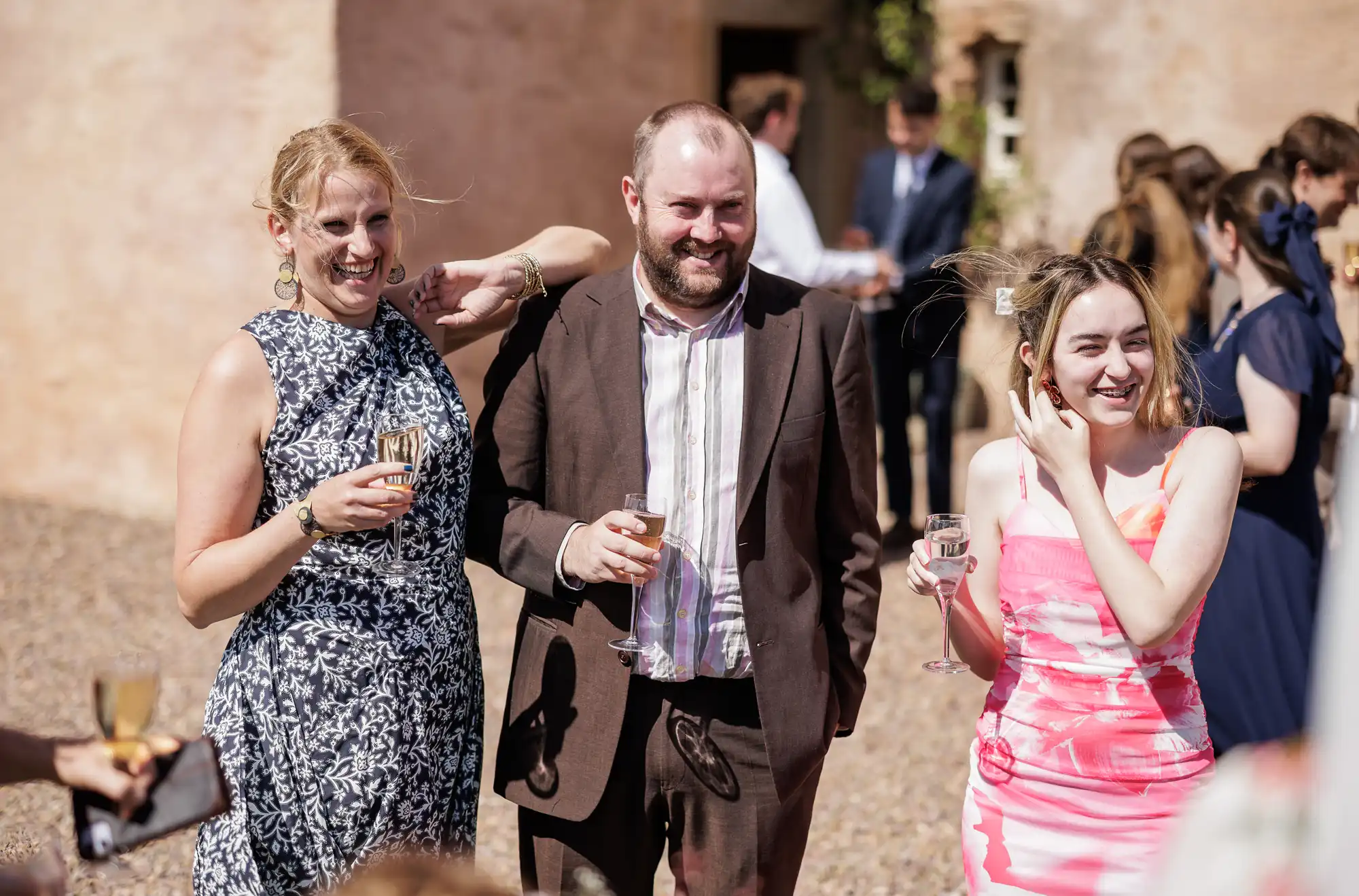 Three people stand outside at a social event, smiling and holding drinks. A woman in a black-and-white dress, a man in a brown suit, and a woman in a pink dress are enjoying themselves. Other guests and a sunlit building are seen in the background.