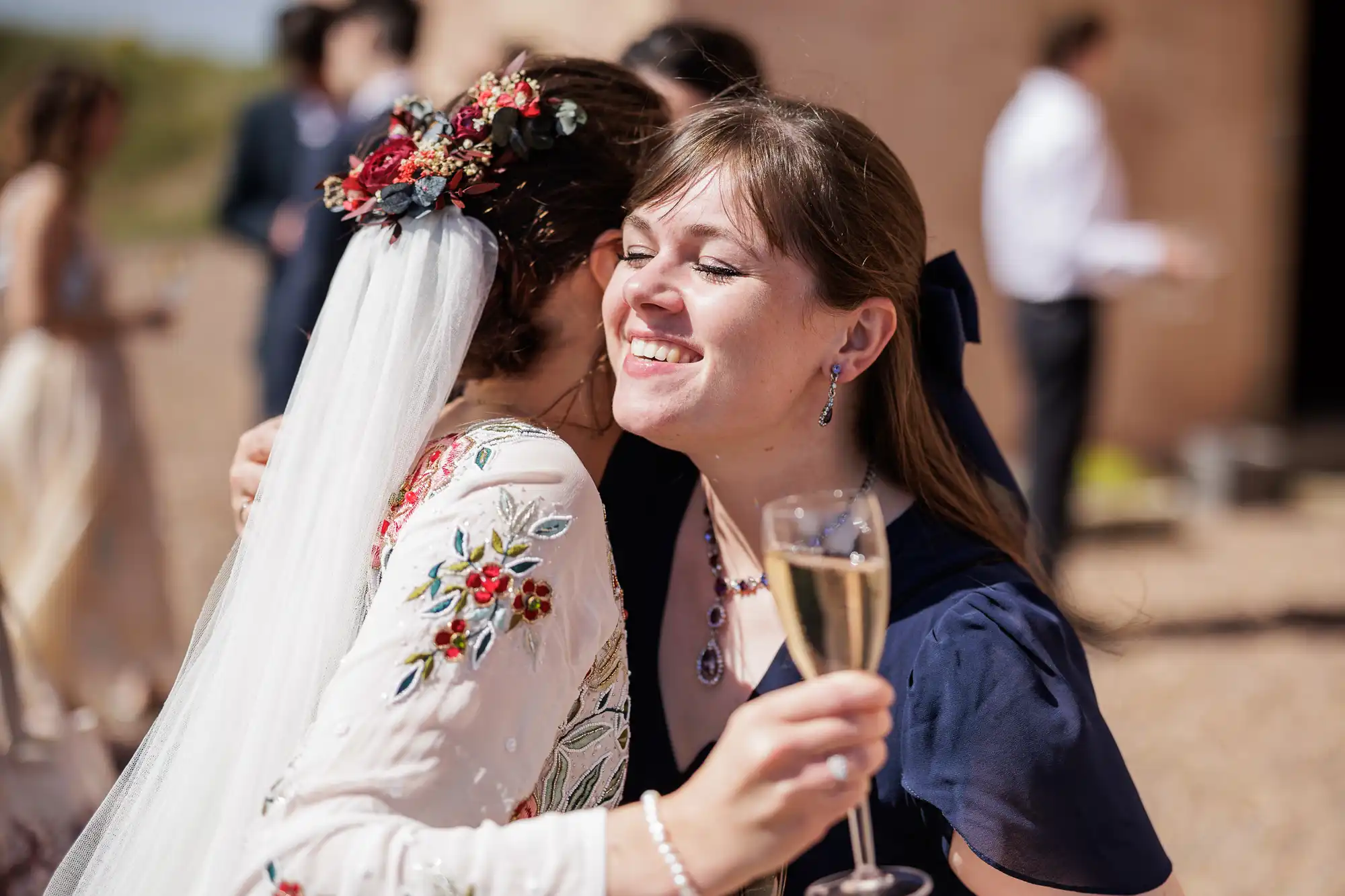 Two women embrace at an outdoor event; one in a floral dress and veil, the other in navy holding a glass of champagne and smiling. The background shows blurred guests and a sunlit stone wall, suggesting a wedding celebration.