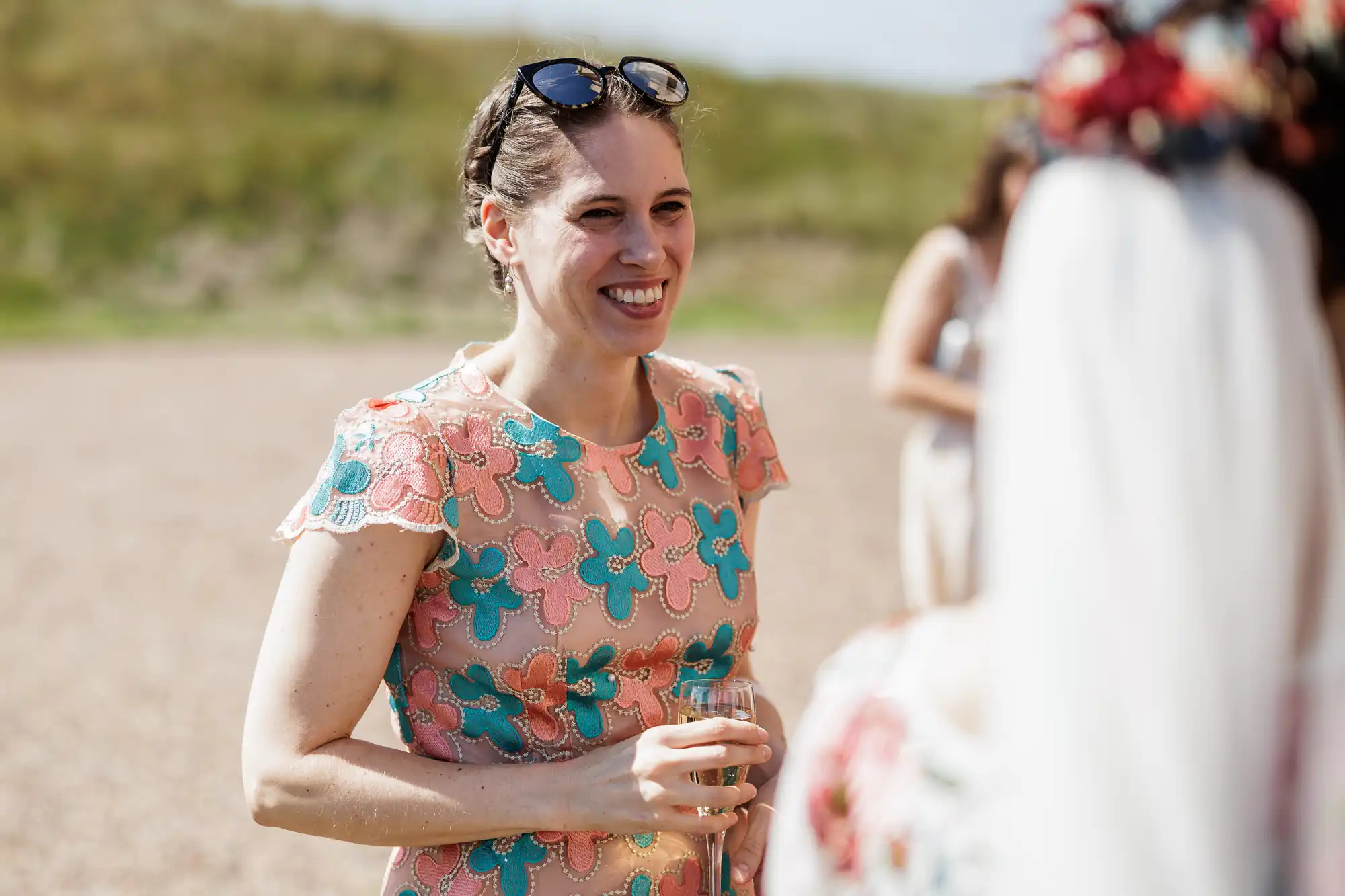 A woman in a peach dress with blue and pink floral patterns smiles while holding a drink. She has sunglasses resting on her head and stands outdoors on a sunny day, talking with another person whose back is to the camera. Blurred grass is in the background.