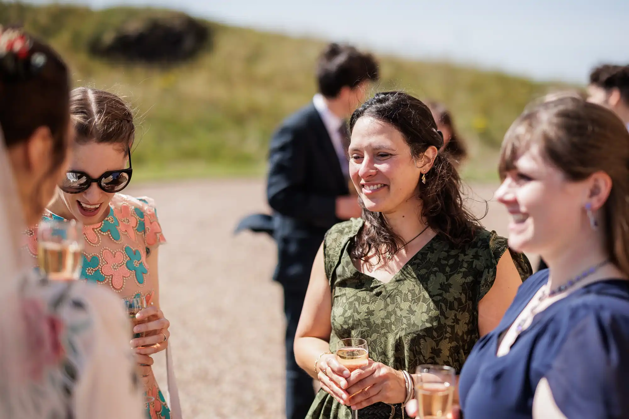 Four women stand outdoors on a sunny day, smiling and holding drinks. They wear colourful dresses and appear to be engaged in conversation. A blurred figure in a suit stands in the background, and grassy hills are visible behind them.