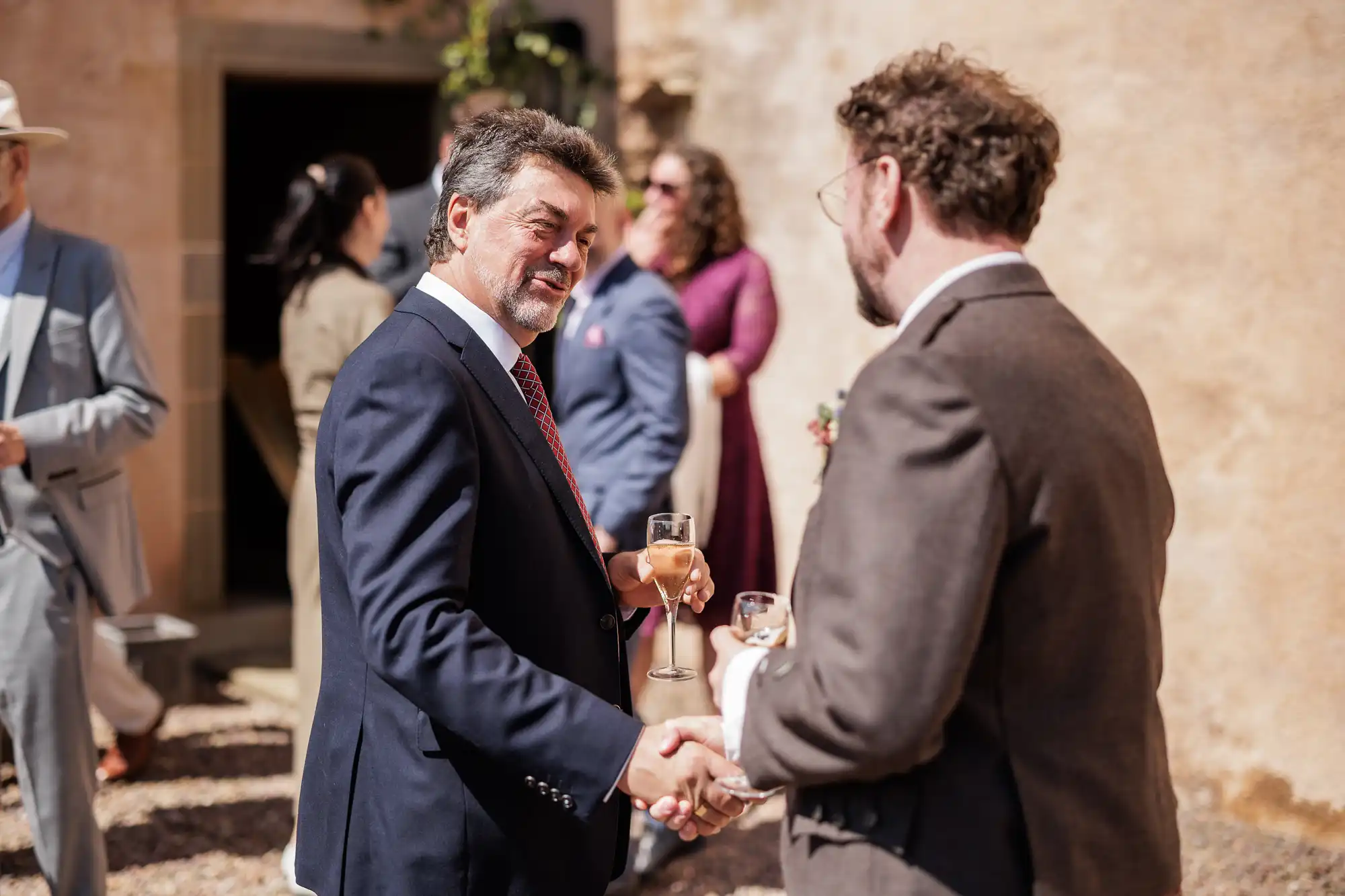 Two men in suits shake hands and smile outdoors at a daytime event. One holds a champagne flute. People in formal attire socialise in the background near a peach-coloured wall. Sunlight creates a cheerful and celebratory atmosphere.