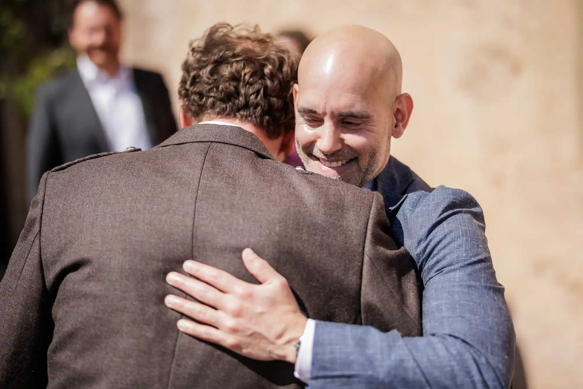 A bald man in a blue suit smiles warmly as he hugs another man in a brown jacket. The focus is on their embrace, showing joy and connection. The background is blurred, with another person visible but out of focus.