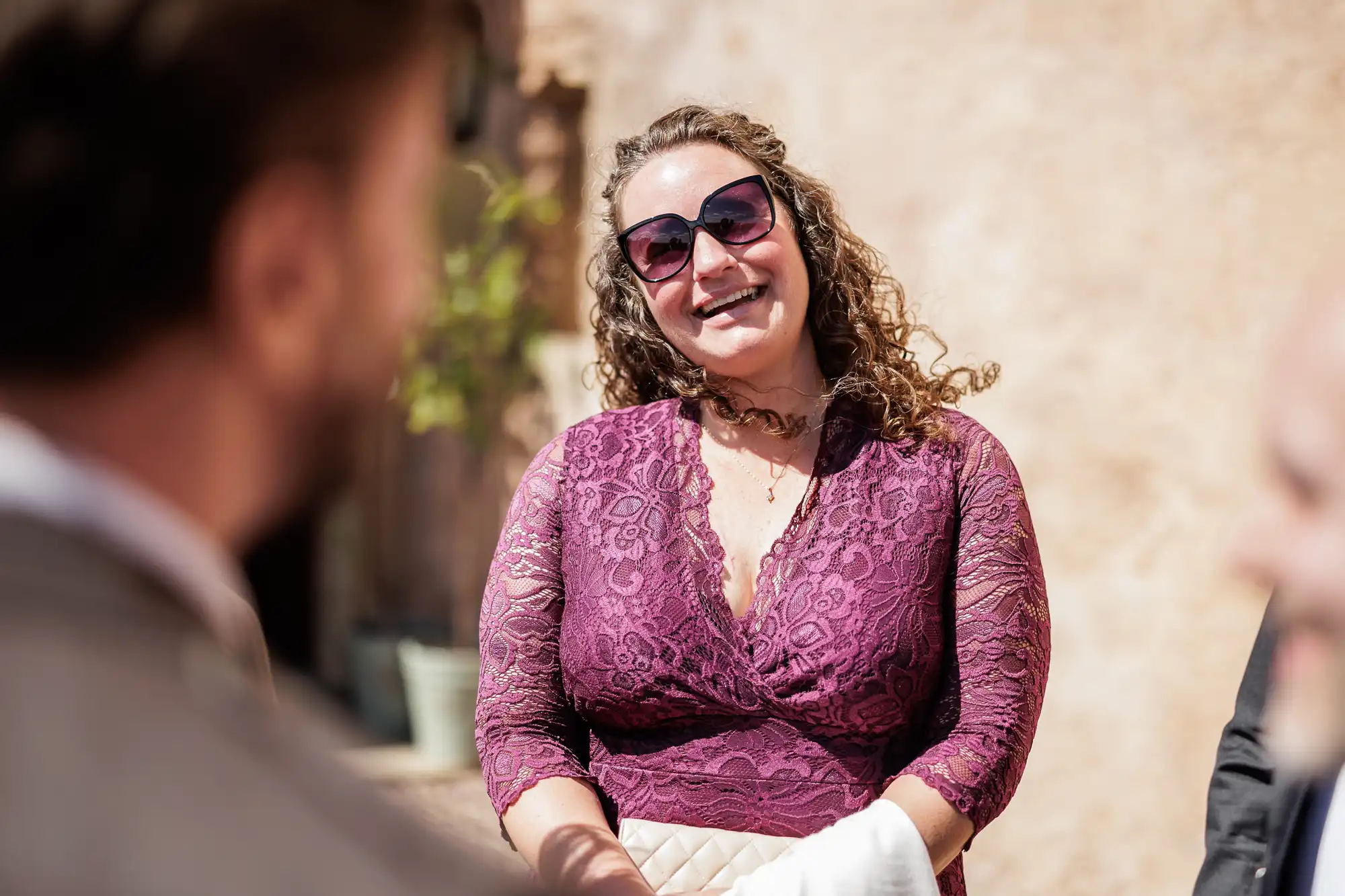 A smiling woman with curly hair, wearing sunglasses and a purple lace dress, stands outdoors in sunlight. She holds a light-coloured clutch and appears to be engaged in conversation with two blurred figures in the foreground.