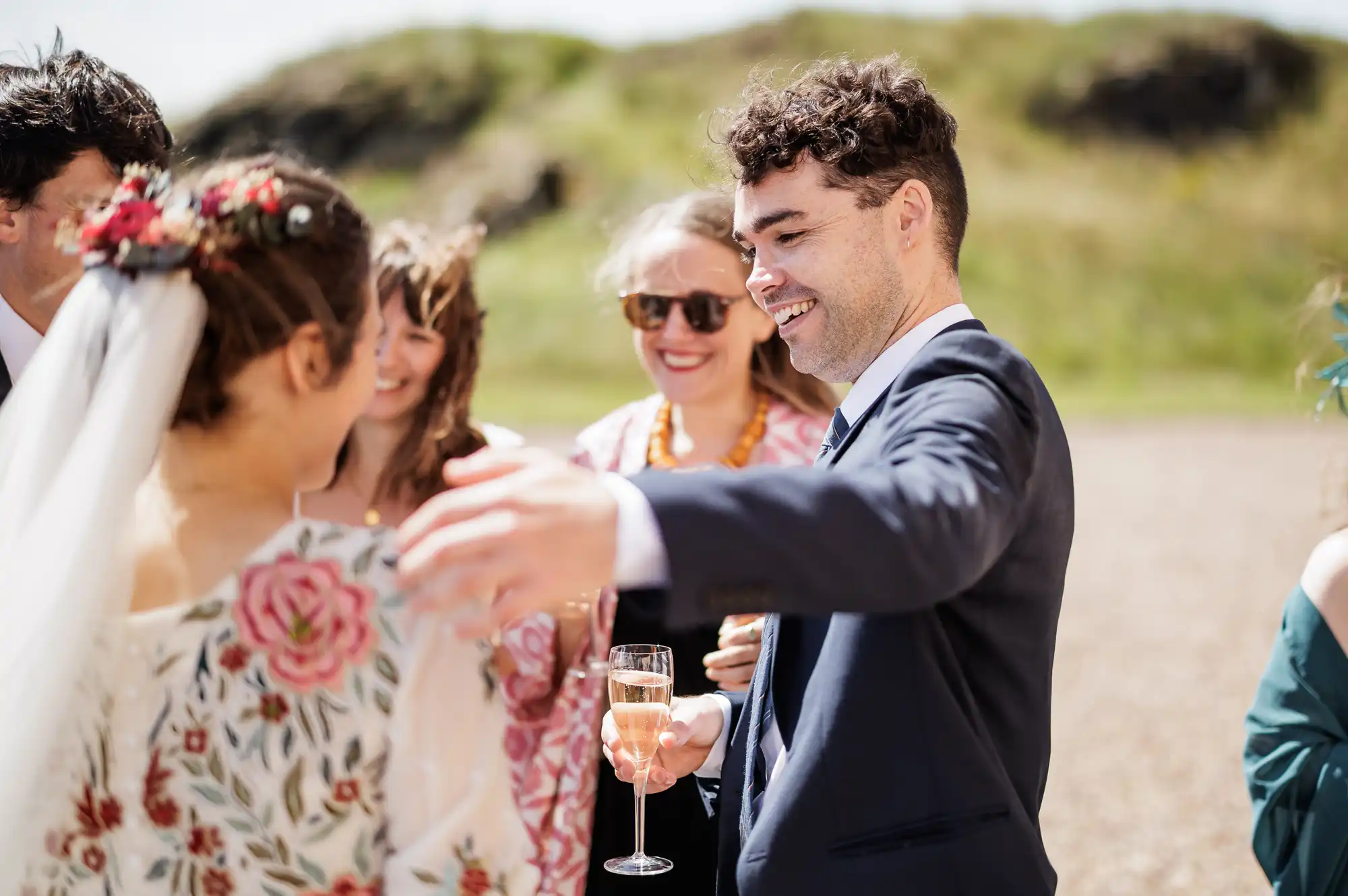 A man in a suit holding a glass of champagne smiles and reaches out to embrace a woman in a white veil with floral embroidery. Several other people, casually dressed and smiling, stand around them outdoors on a sunny, grassy day.