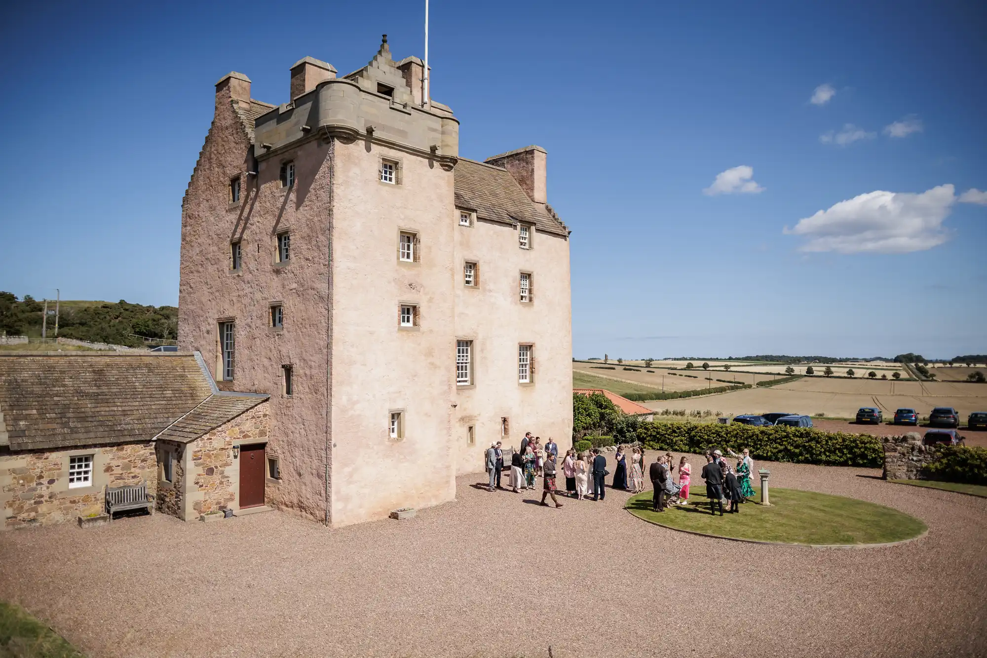 A group of people gathers outside a tall, stone castle with small windows, set in a rural landscape with fields, a clear sky, and parked cars in the distance. The scene is bright and sunny, suggesting a special event or gathering.