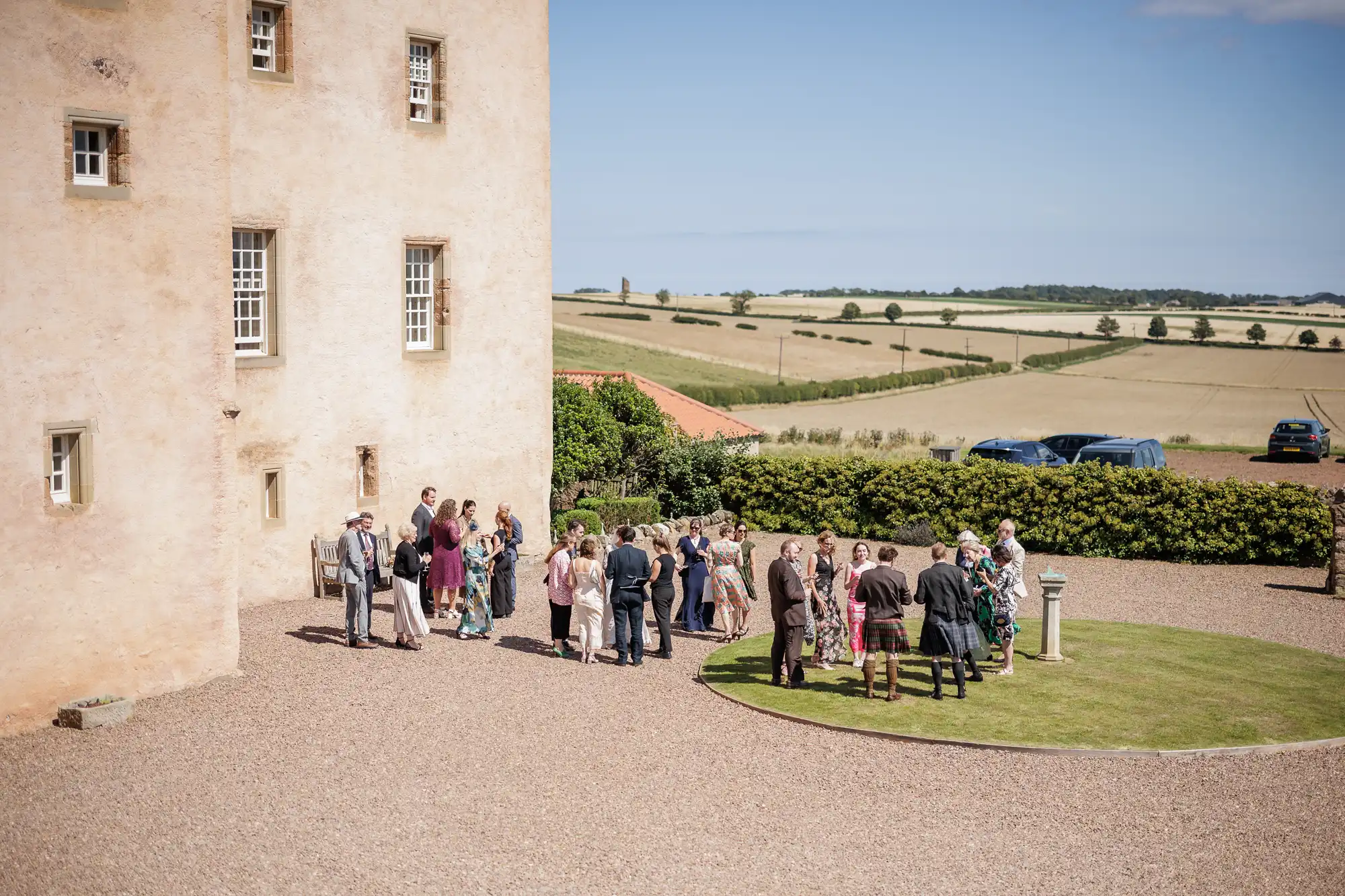 A group of people gather on a gravel path and small grassy circle near a large, pale stone building in the countryside. Cars are parked by a hedge, and fields stretch into the distance under a blue sky with scattered clouds.