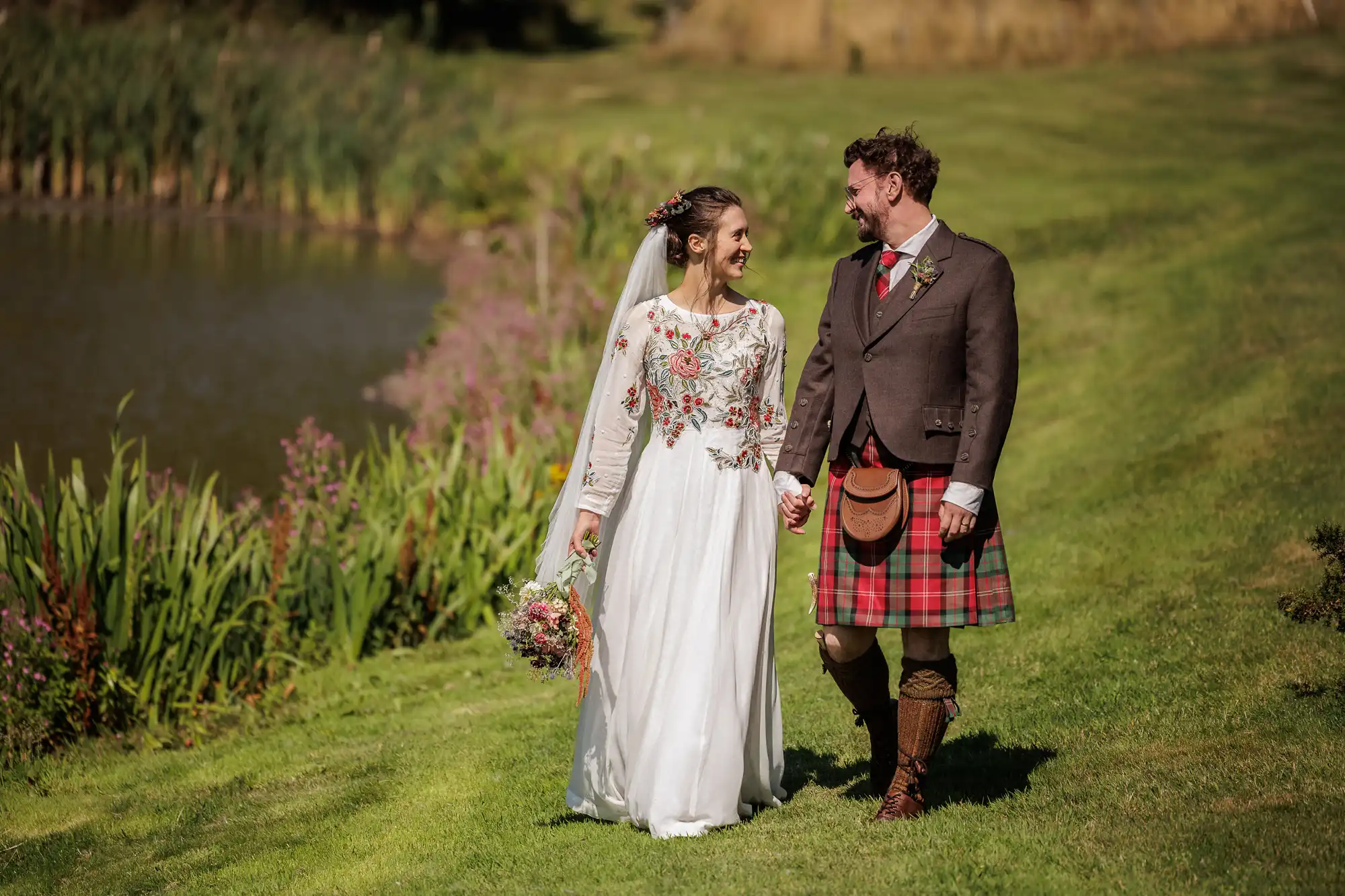 A bride in a white dress with floral embroidery and a groom in a brown jacket with a red tartan kilt walk hand-in-hand on green grass, smiling at each other near a pond and colourful wildflowers on a sunny day.