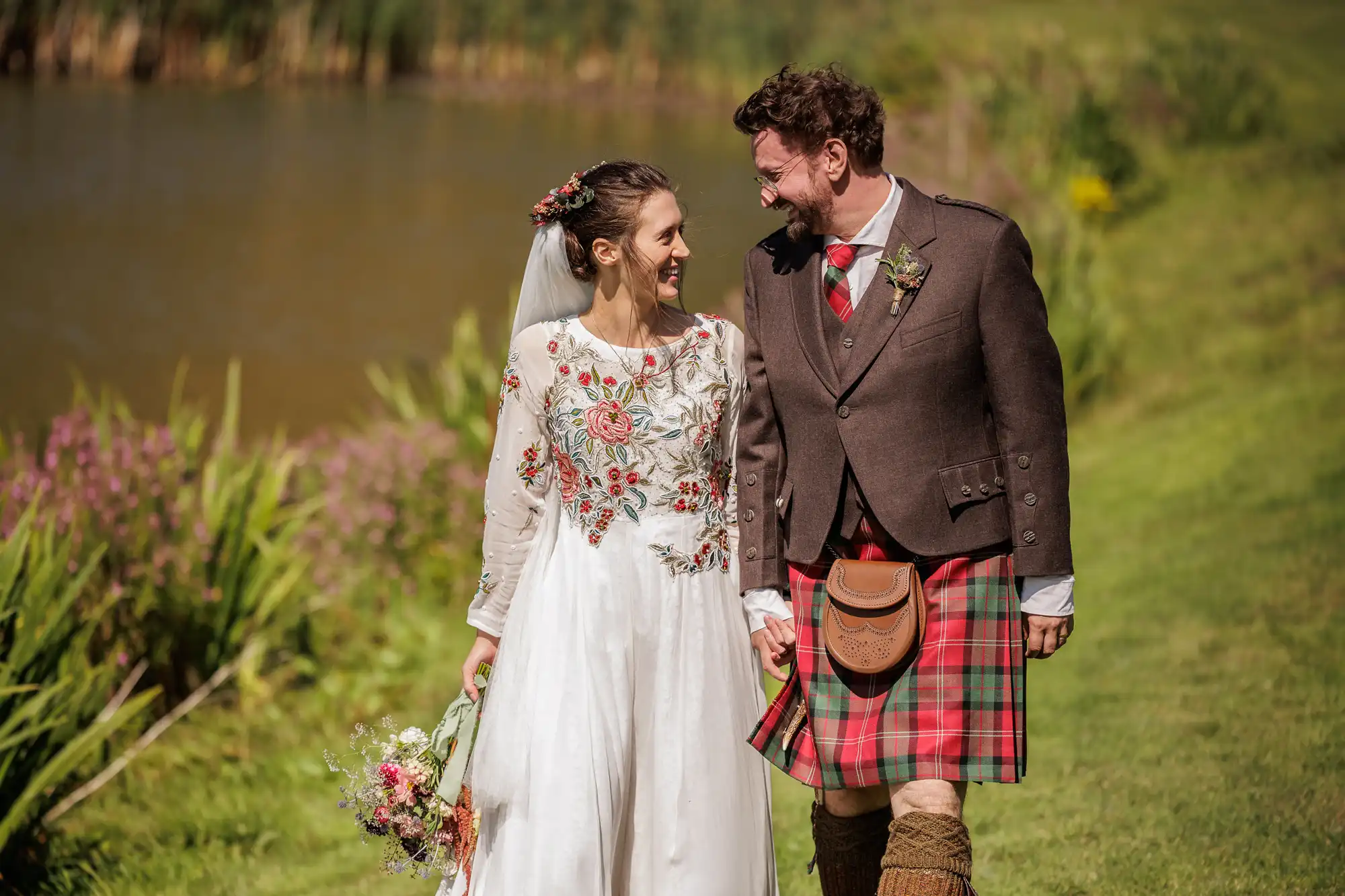 A smiling bride in a white, floral-embroidered dress holding a bouquet walks hand-in-hand with a groom in a brown jacket and red checked kilt. They gaze at each other, standing on green grass by a pond, with wild flowers and bright sunlight around them.