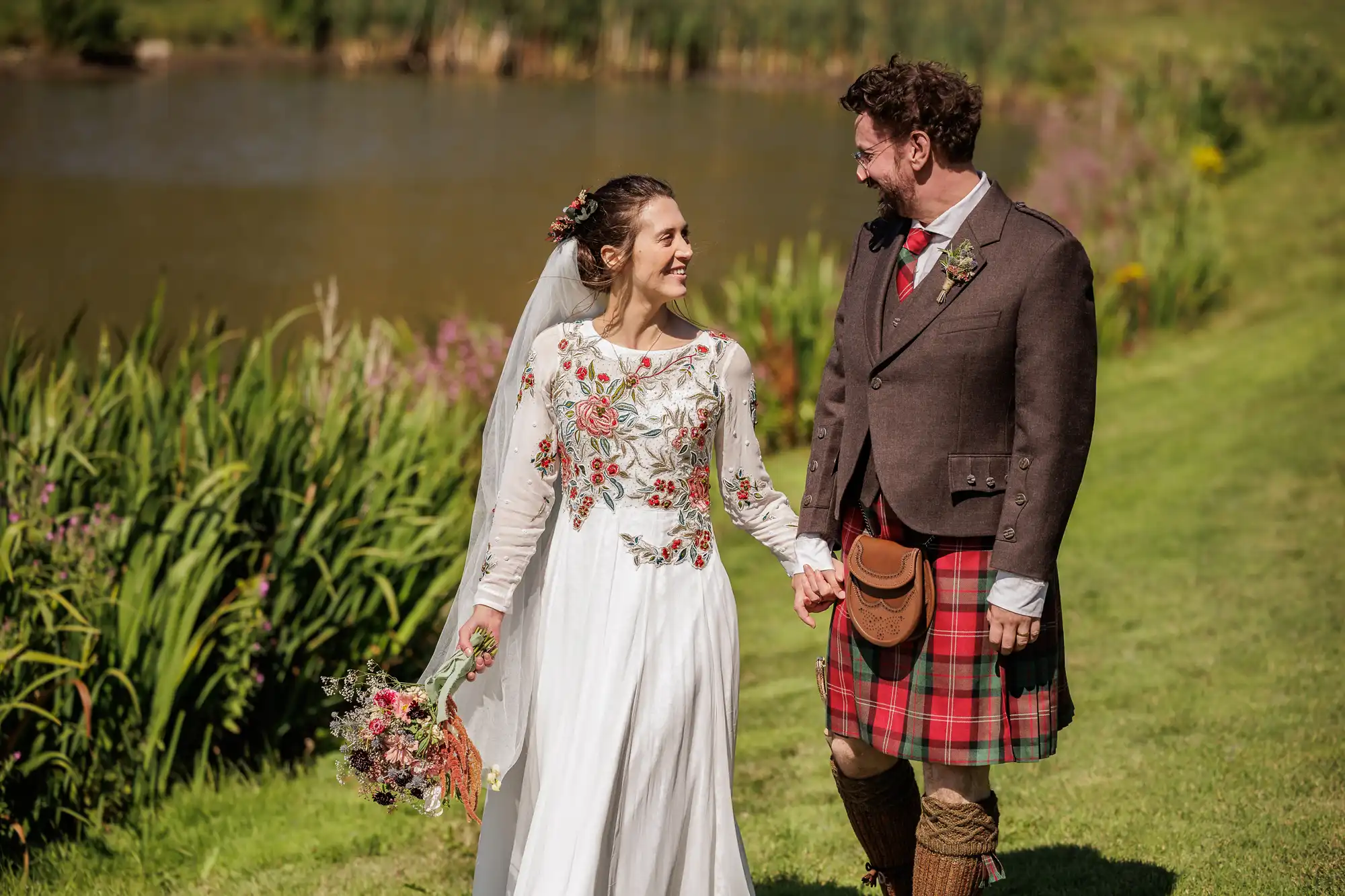 A smiling bride in a long white floral dress and a groom in a brown jacket with a red tartan kilt walk hand-in-hand on grass by a pond, with greenery and flowers around them. The bride holds a bouquet and wears a veil. Both look happy and gaze at each other.