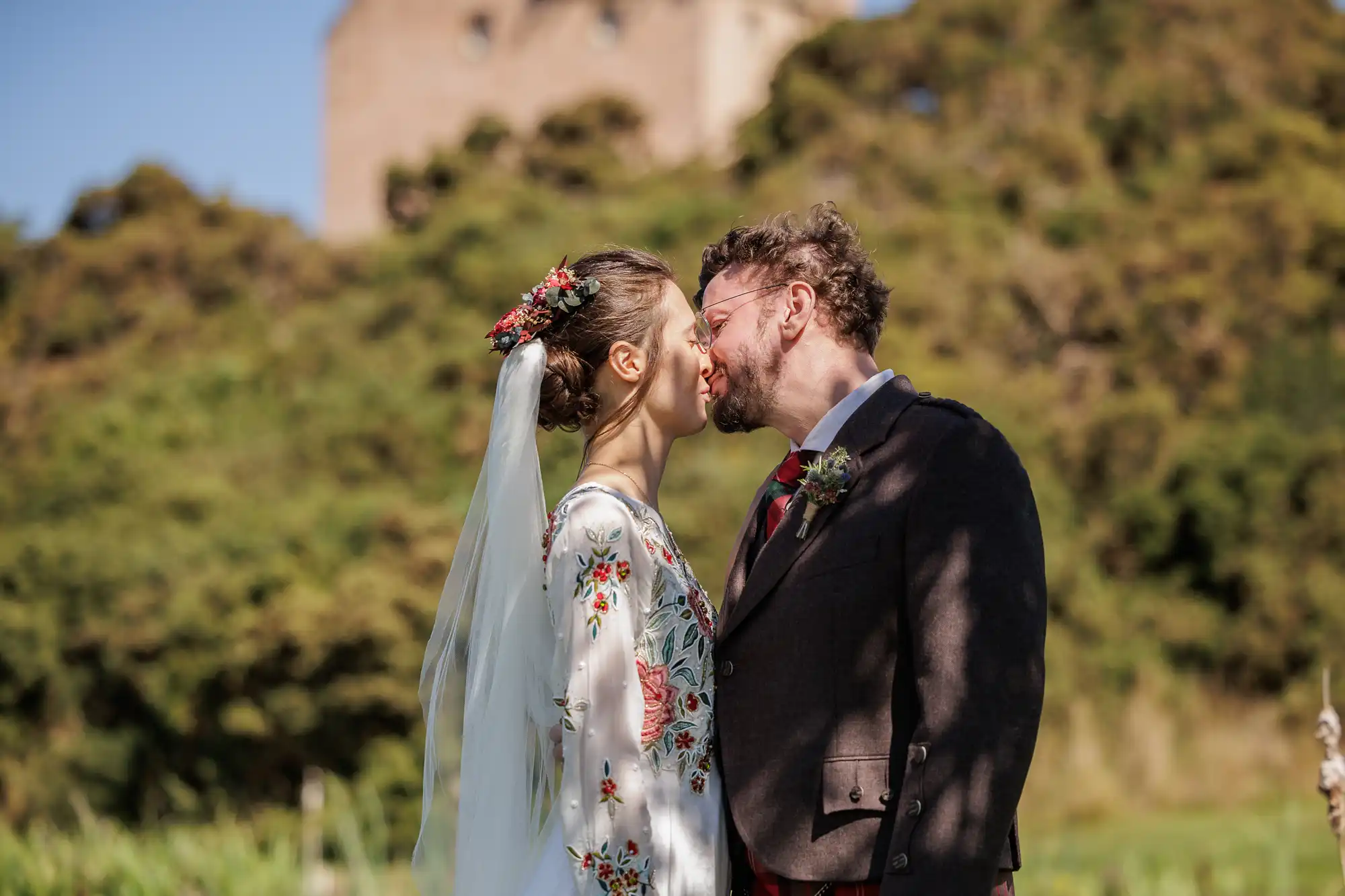 A bride and groom stand outdoors, gently touching noses and smiling. The bride wears a floral embroidered dress with a long veil and flowers in her hair. The groom wears glasses, a dark jacket, and a buttonhole. Green trees and a blurred stone building are in the background.