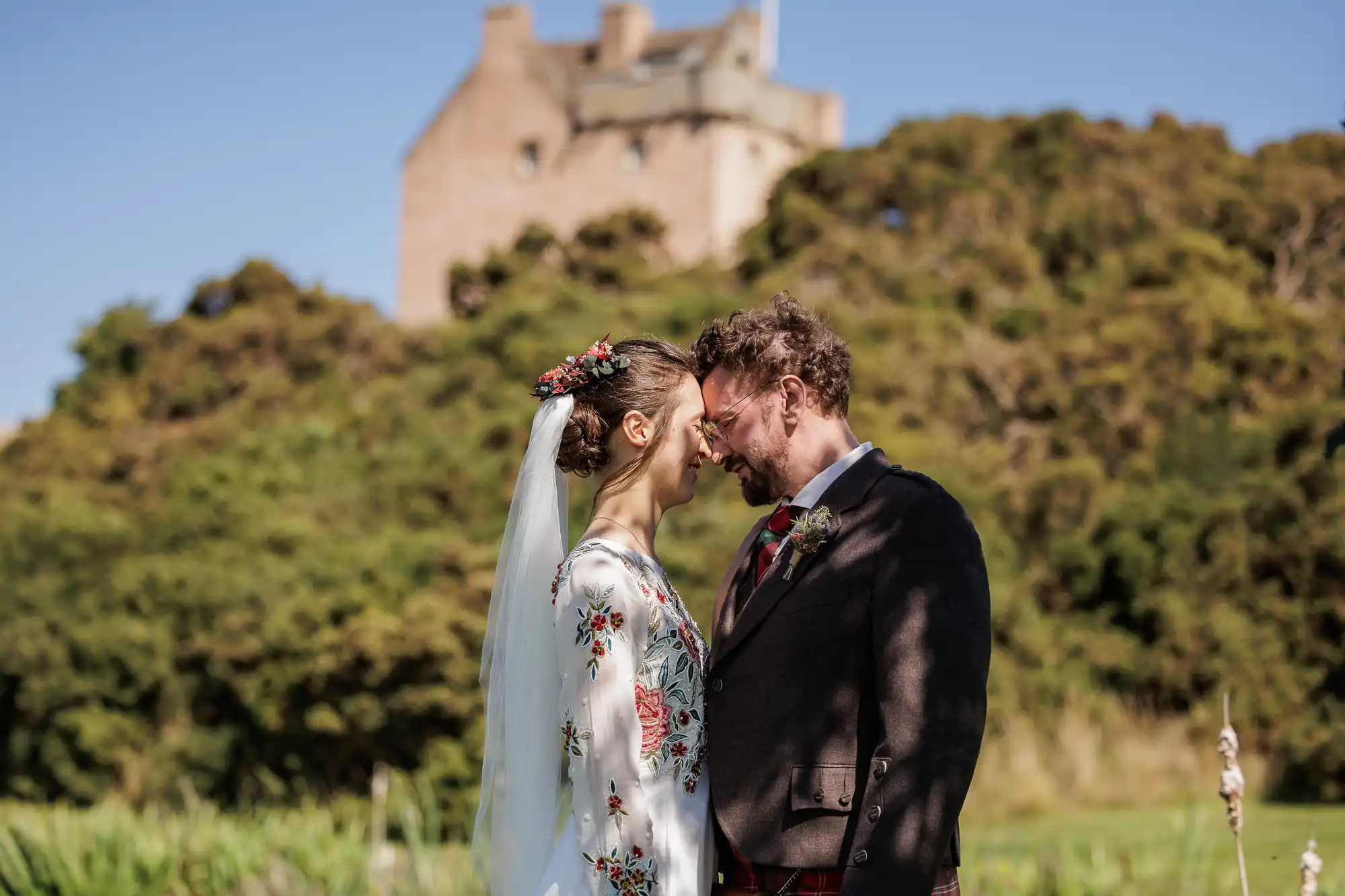 A bride in a white dress with floral embroidery and a veil and a groom in a dark suit with a tartan tie stand outdoors, smiling with their foreheads touching. Behind them are greenery and a large stone building on a hill under a blue sky.