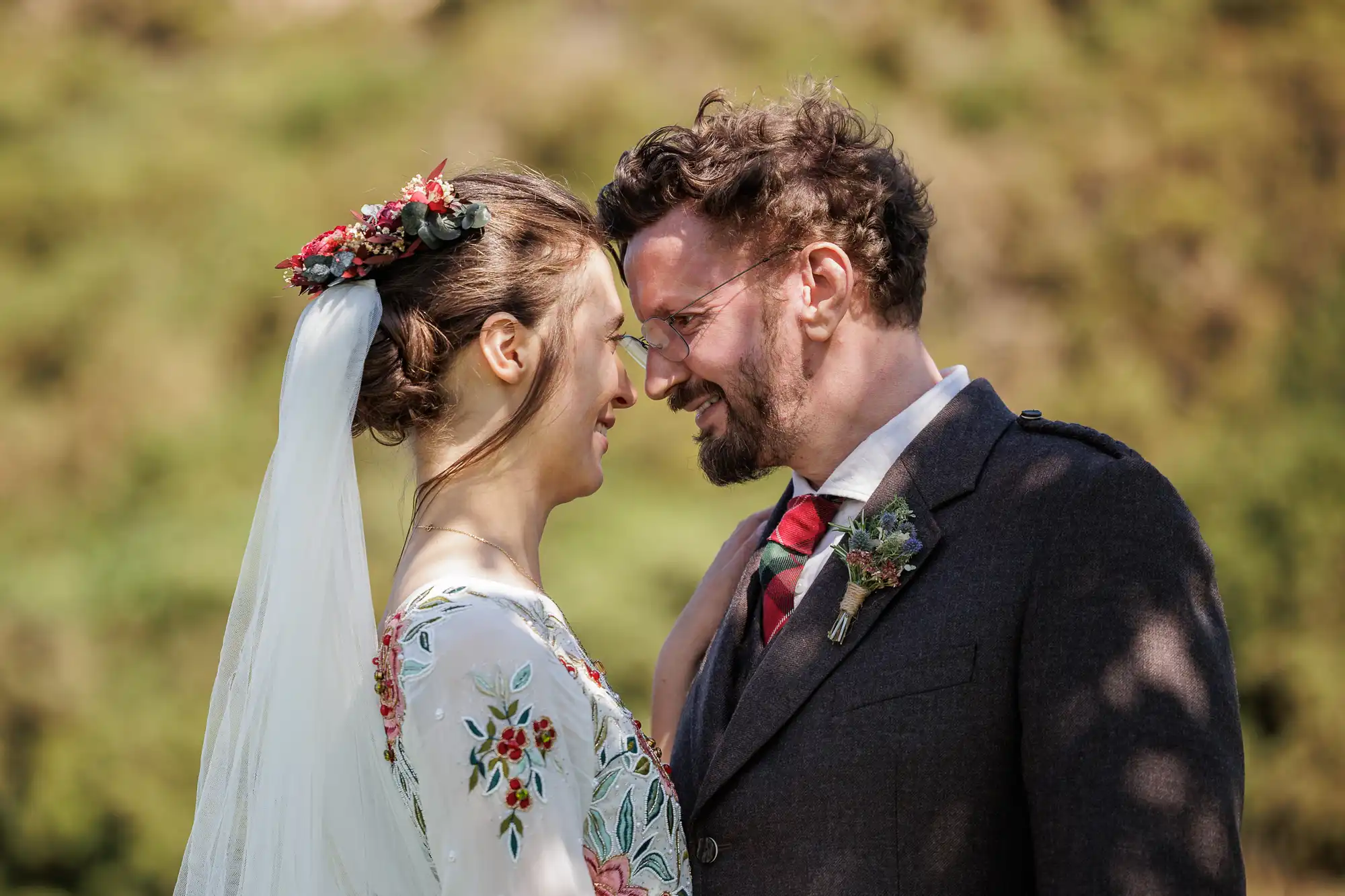 A smiling bride and groom stand close together, touching foreheads. The bride wears a white dress with colourful floral embroidery and a veil with flower accents. The groom wears glasses, a brown suit, a checked tie, and a buttonhole. They are outdoors in sunlight.