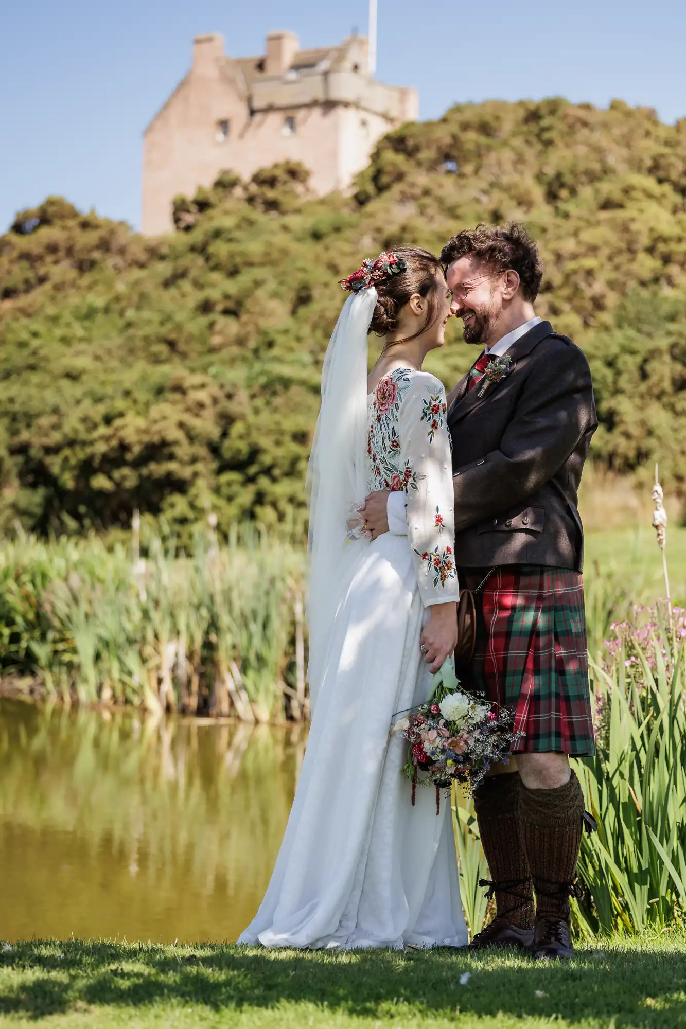 A bride in a long white dress with floral embroidery and a groom in a Scottish kilt stand embracing beside a pond, smiling at each other, with greenery and a castle in the background under a sunny sky.