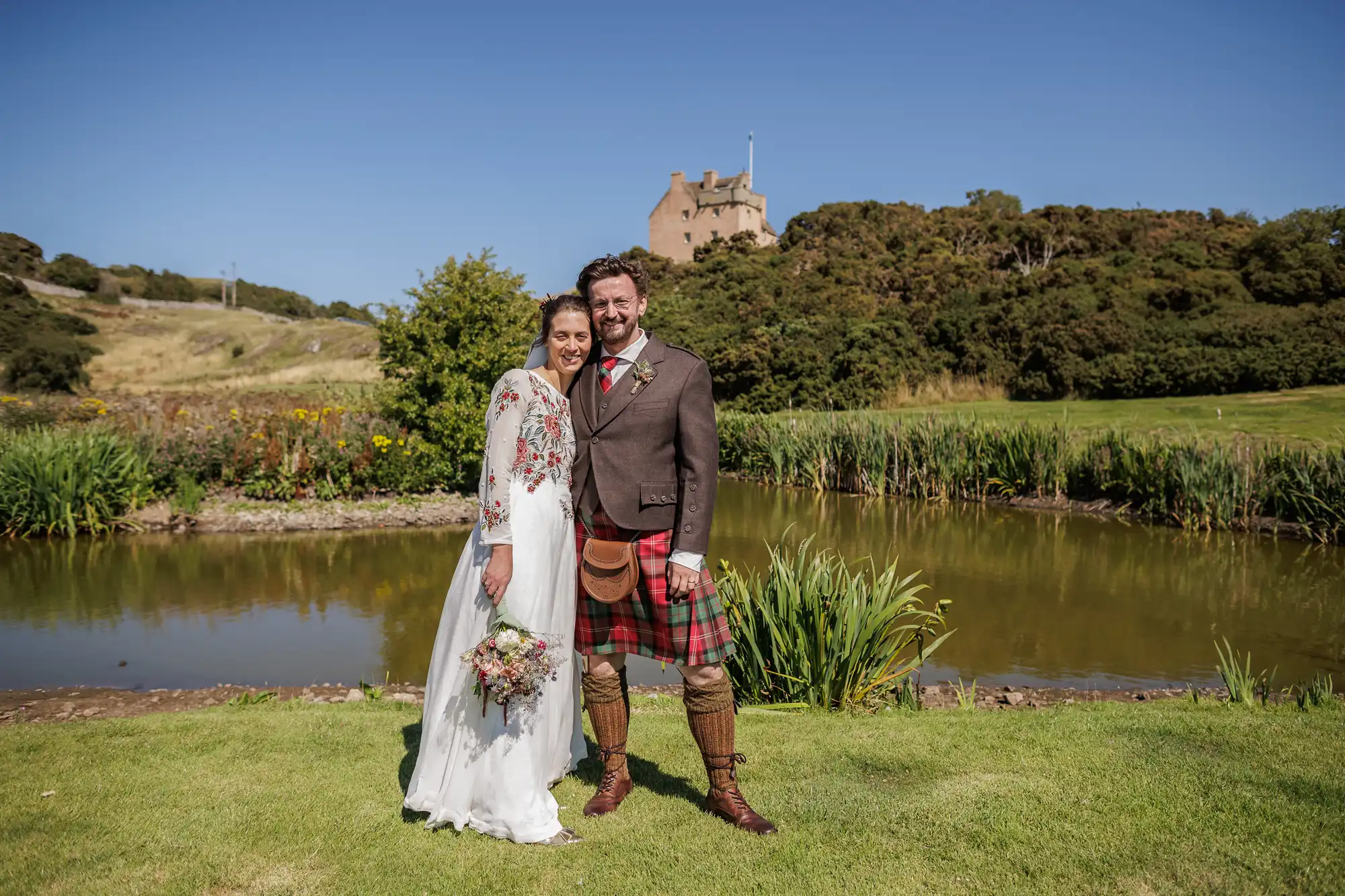 A smiling bride in a white dress and floral shawl stands beside a groom in a brown jacket, kilt, and sporran. They pose outdoors by a pond with wild flowers. Green hills and a castle atop a wooded hill appear in the background under a clear blue sky.
