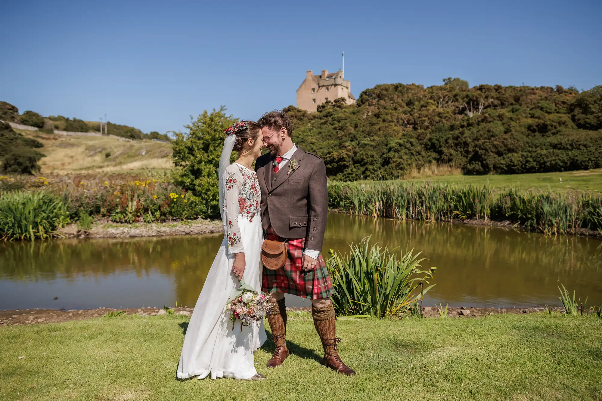 A bride in a white floral dress and veil stands forehead-to-forehead with a groom in a dark jacket and red tartan kilt by a pond, with lush greenery and a stone castle on a hill in the background under a clear blue sky.